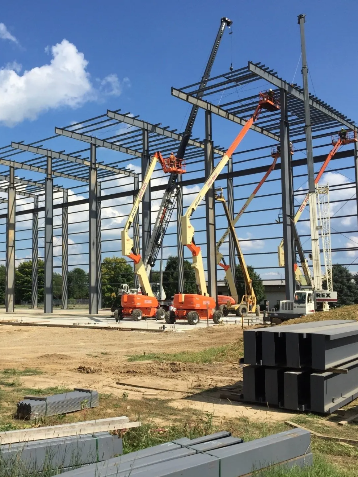 Construction site with multiple cranes lifting steel beams into place for a building framework under a blue sky with clouds.