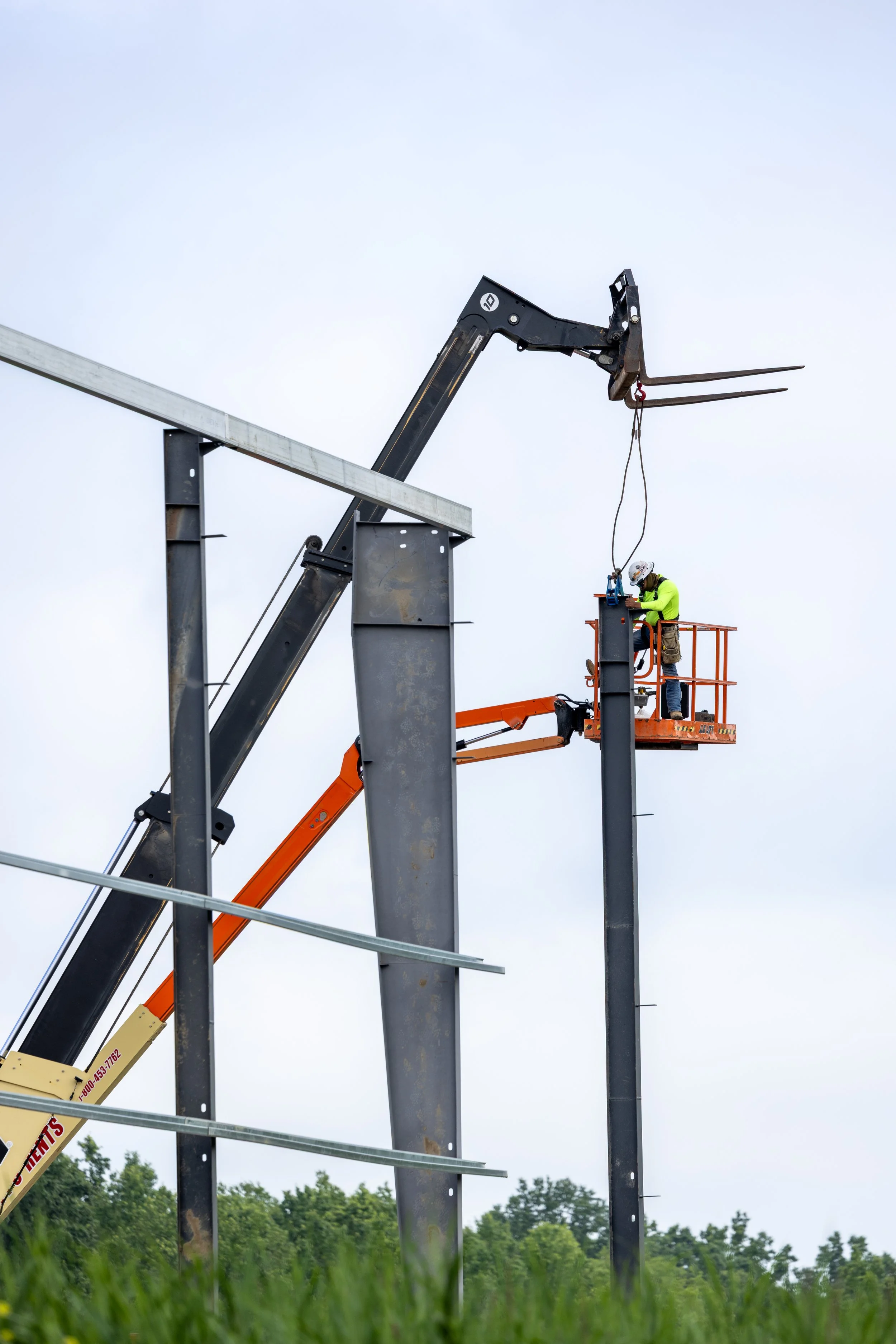 Construction worker in safety gear is on an orange lift, working on steel support beams at a construction site of a large structure, with a cloudy sky and trees in the background.