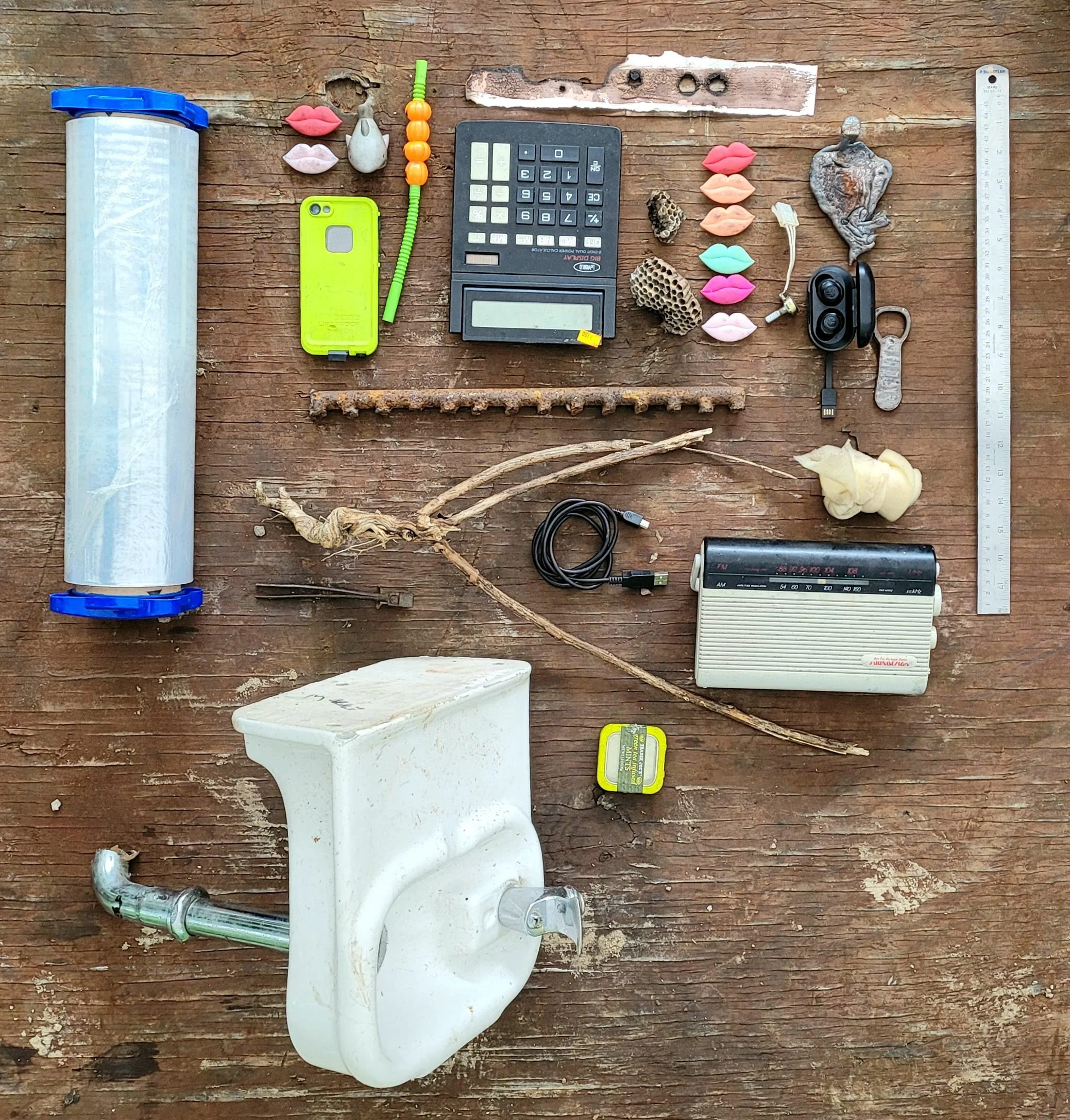 Various objects arranged on a wooden surface including a large plastic roll, lips-shaped objects, small stones, a calculator, a bright yellow phone case, seashells, a mini fan, a memory card, sunglasses, a ruler, and small sticks or branches.