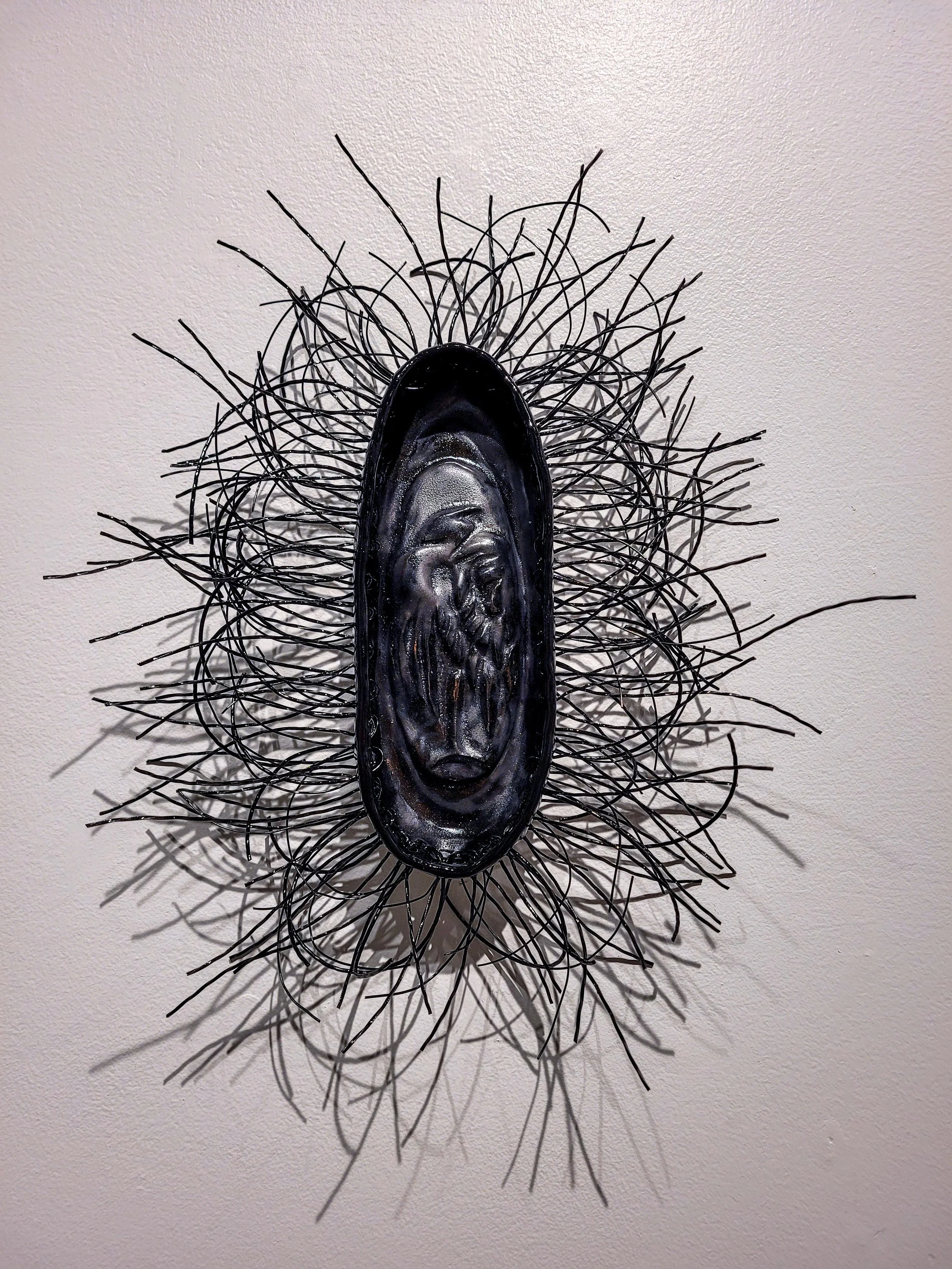 Decorative wall art consisting of a black oval shape with a face in the center, surrounded by radiating black wire extensions, mounted on a white wall.
