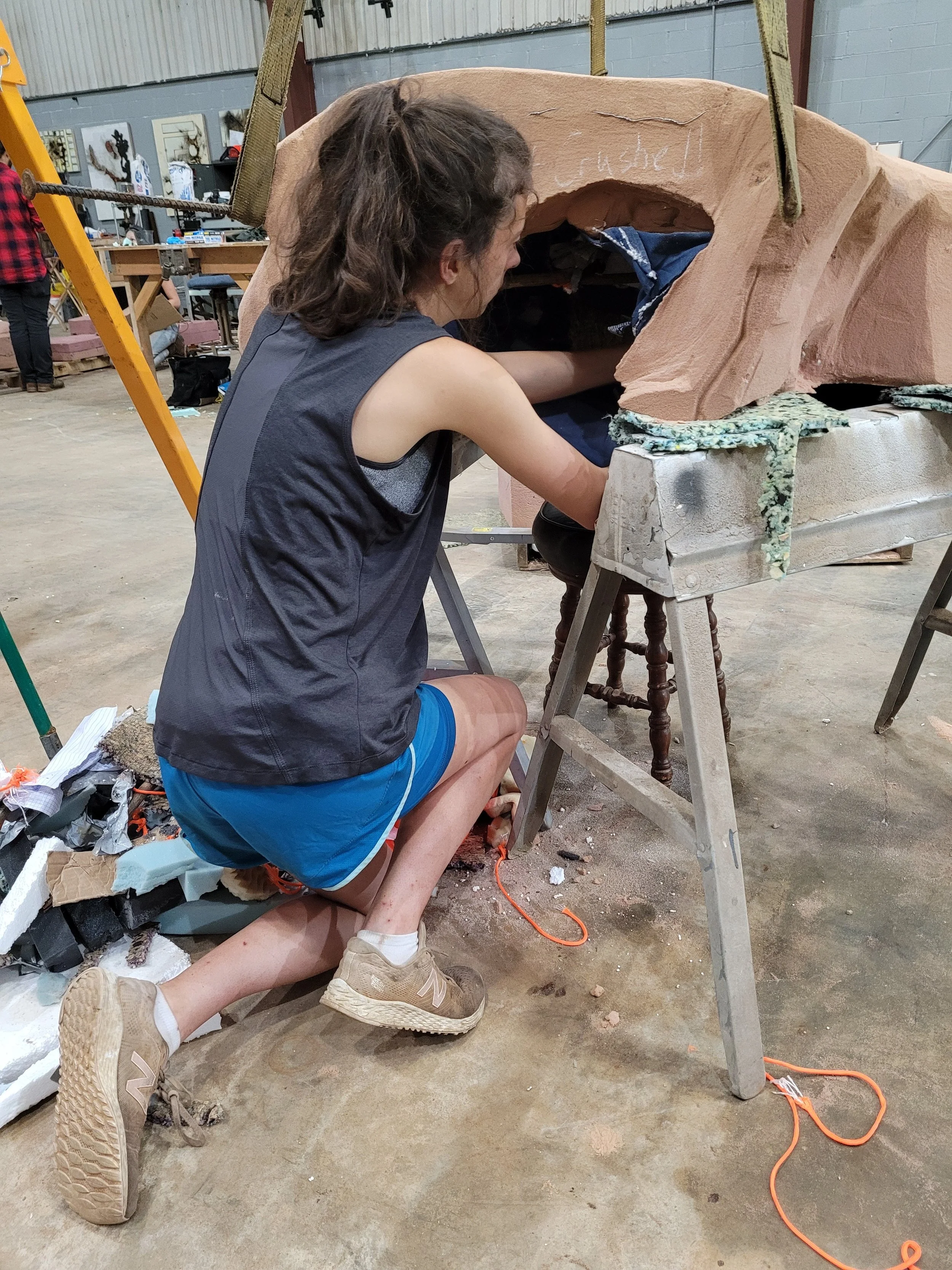 A person crouching down inside a suspended sand mold, working on a project in an industrial workspace with tools and art pieces in the background.