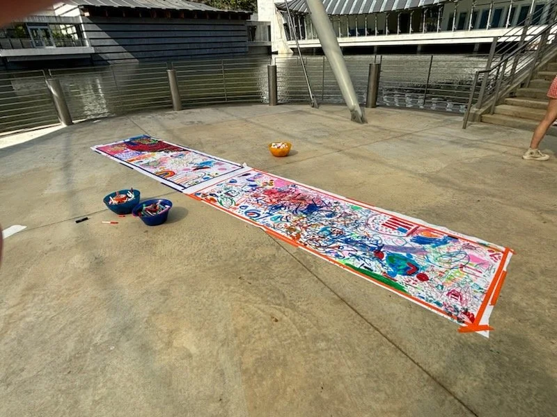 A long stretch of butcher paper taped on the ground covered in colorful child drawings and scribbles with bowls of markers nearby, outdoors near a body of water and modern buildings.