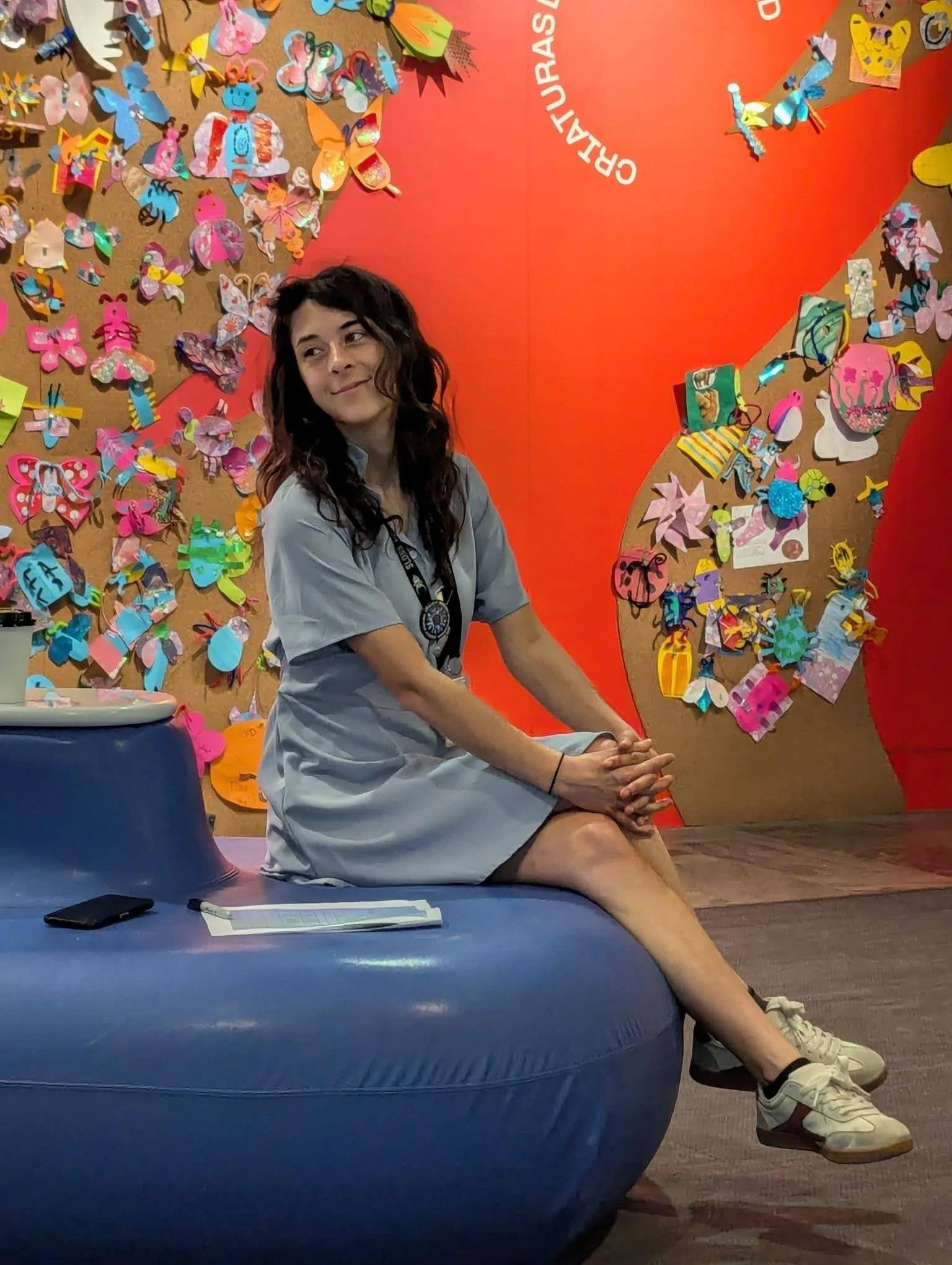 A woman sitting on a blue chair inside an exhibit with colorful paper crafts on a brown, corkboard background. The woman is wearing a gray shirt and shorts, with white sneakers, and has dark wavy hair.