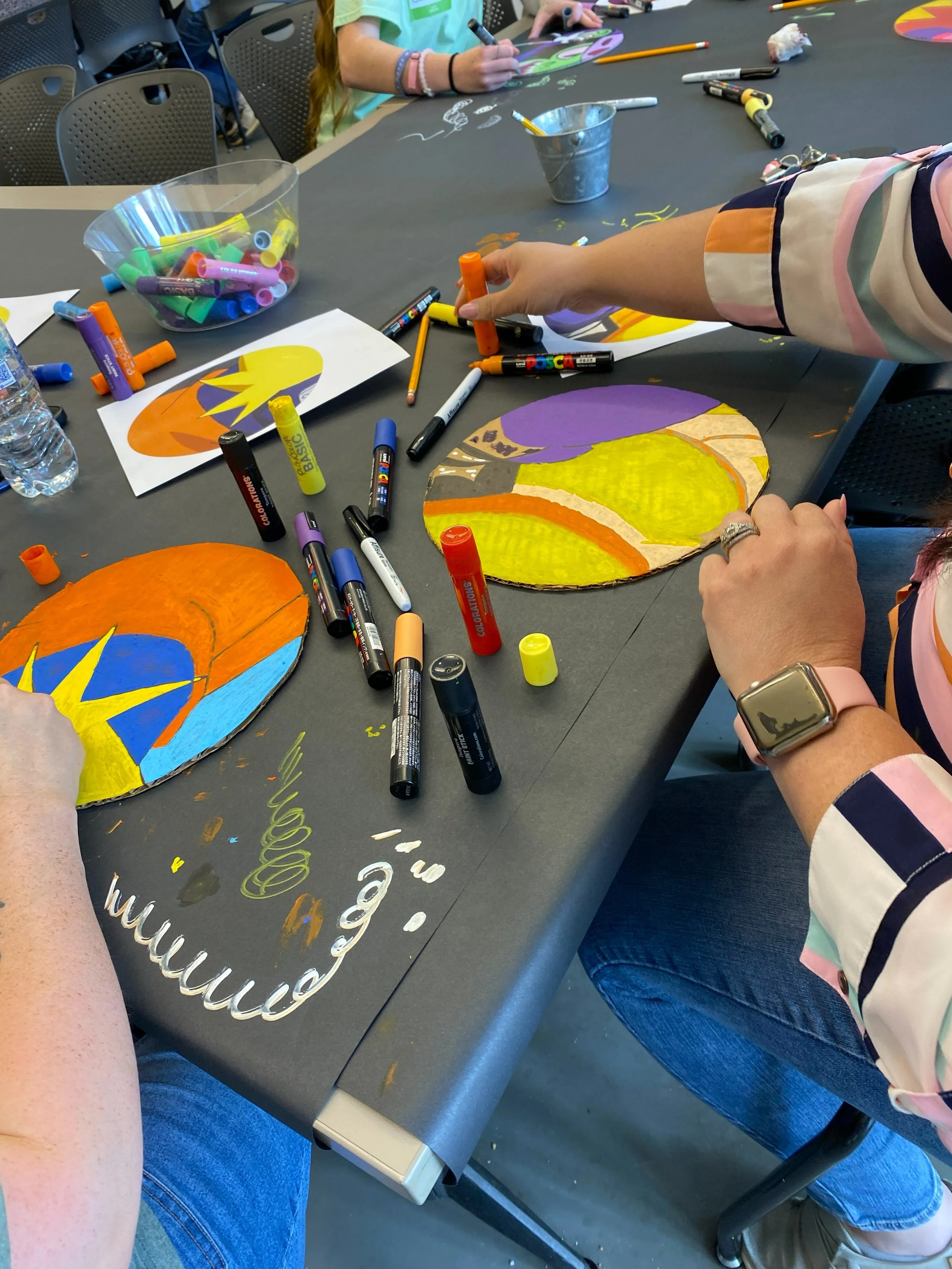 People sitting around a table engaged in arts and crafts, including paint, markers, and painted paper plates with colorful designs. There is a bowl of colorful markers, a water bottle, and various art supplies scattered on the table.