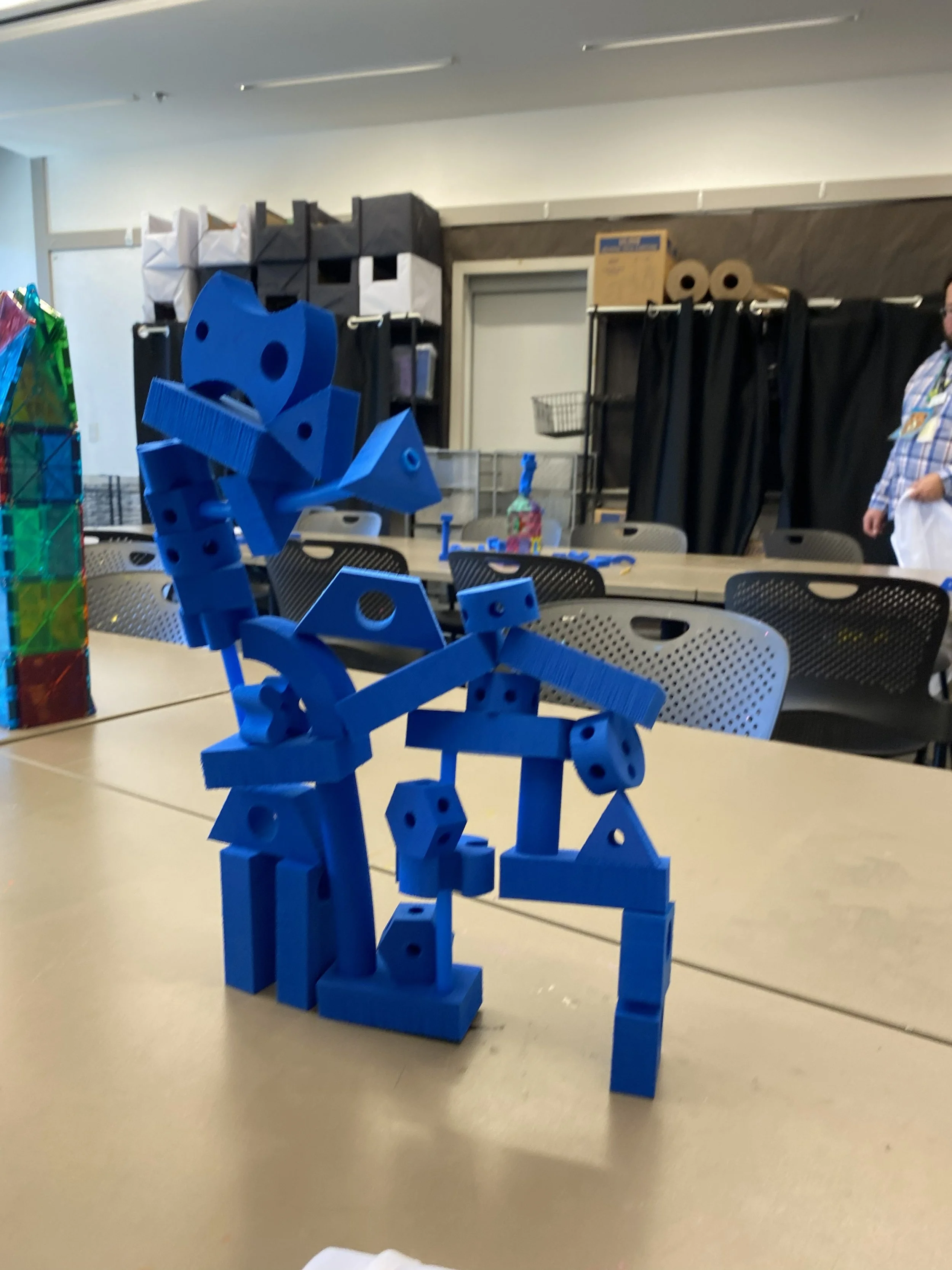 A child's blue abstract sculpture constructed from foam building blocks standing on a beige table in a room with black curtains and shelving in the background.
