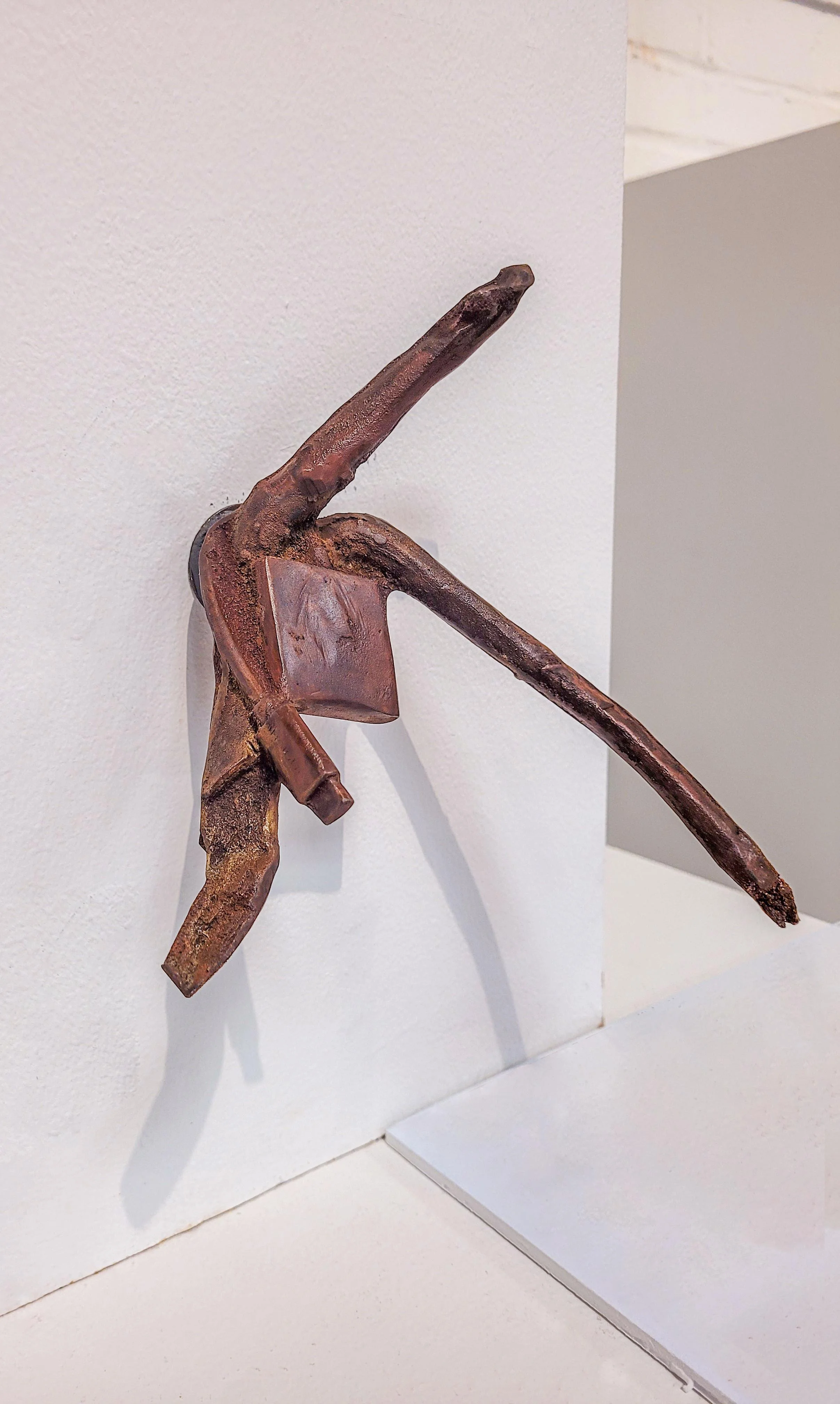 Rusty metal sculpture resembling a human figure with elongated limbs, mounted on a white wall.