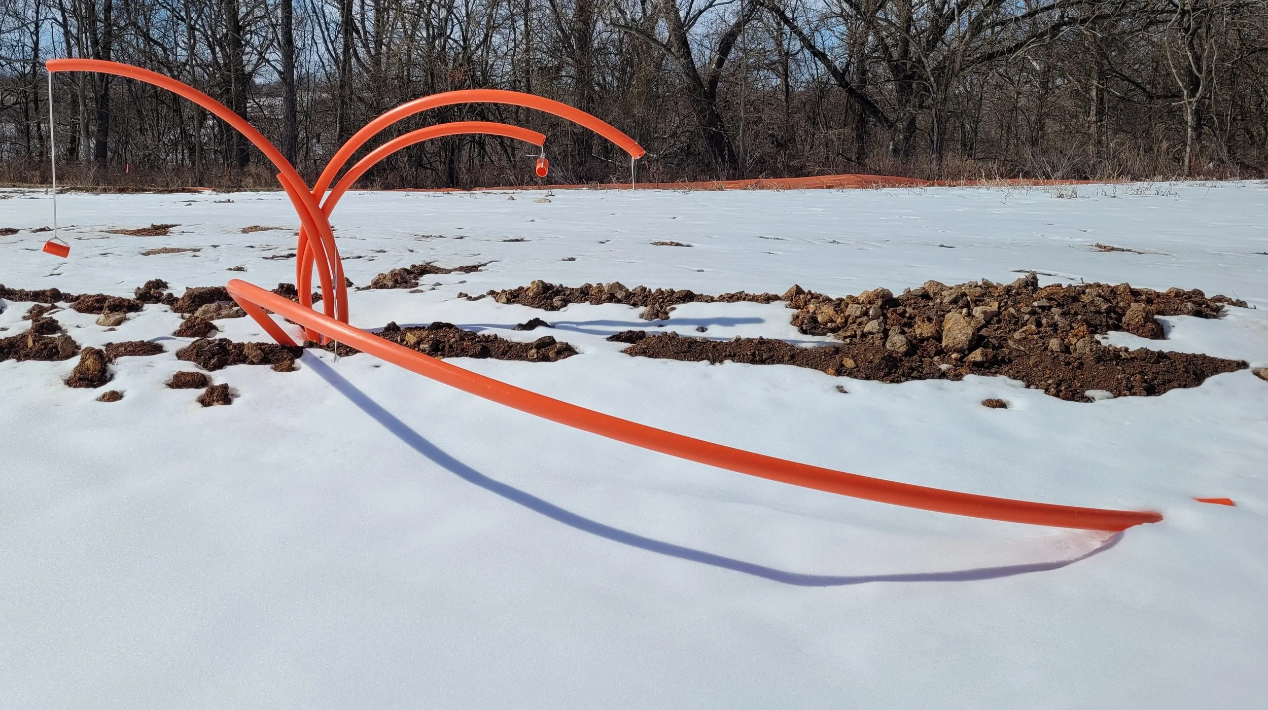 Orange construction pipe on snow-covered ground with dirt patches and a line of leafless trees in the background.