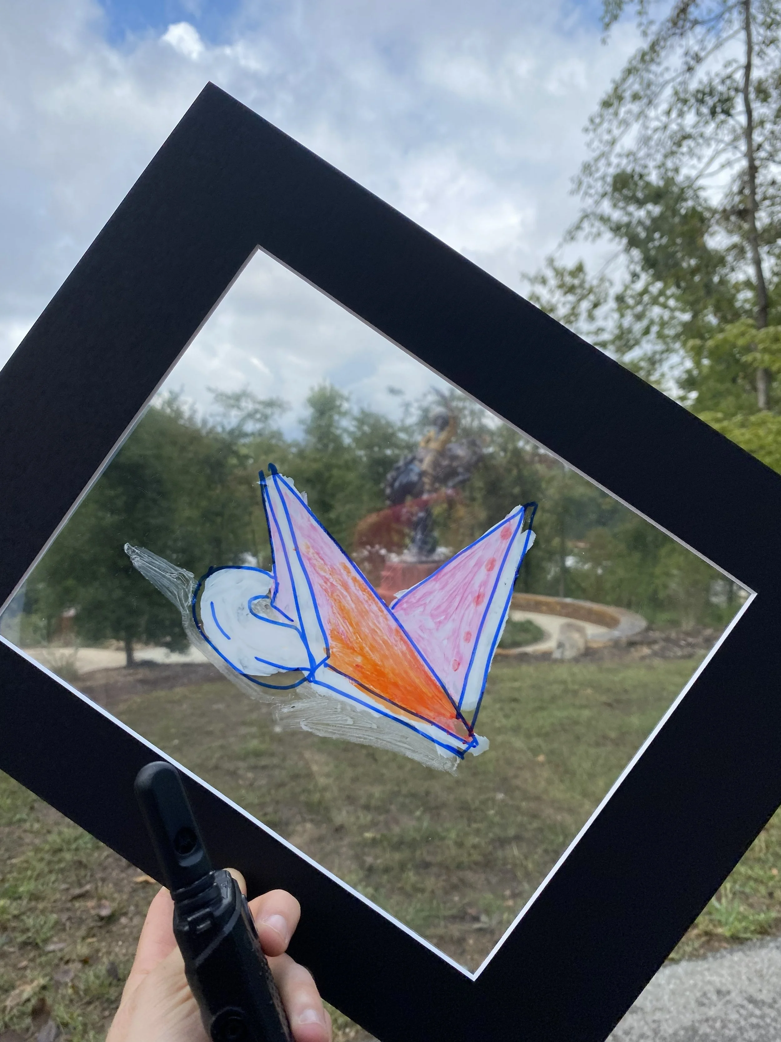 A person holding a mat board framed transparency sheet with a picture of a colorful abstract sculpture drawn on it, through the transparency sheet are the outdoors with trees and a cloudy sky in the background.