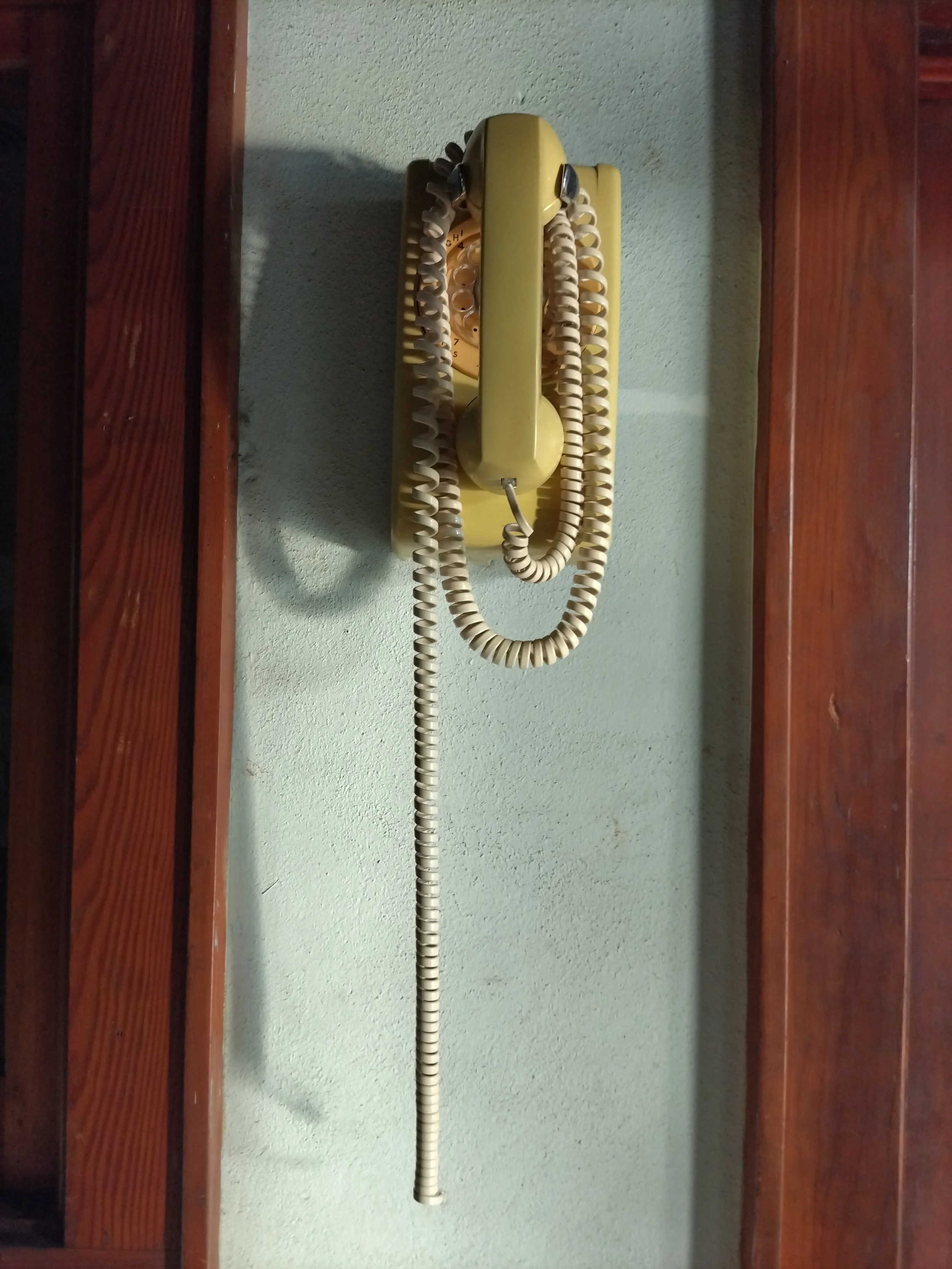 Vintage yellow rotary dial telephone mounted on a green wall between wooden panels.