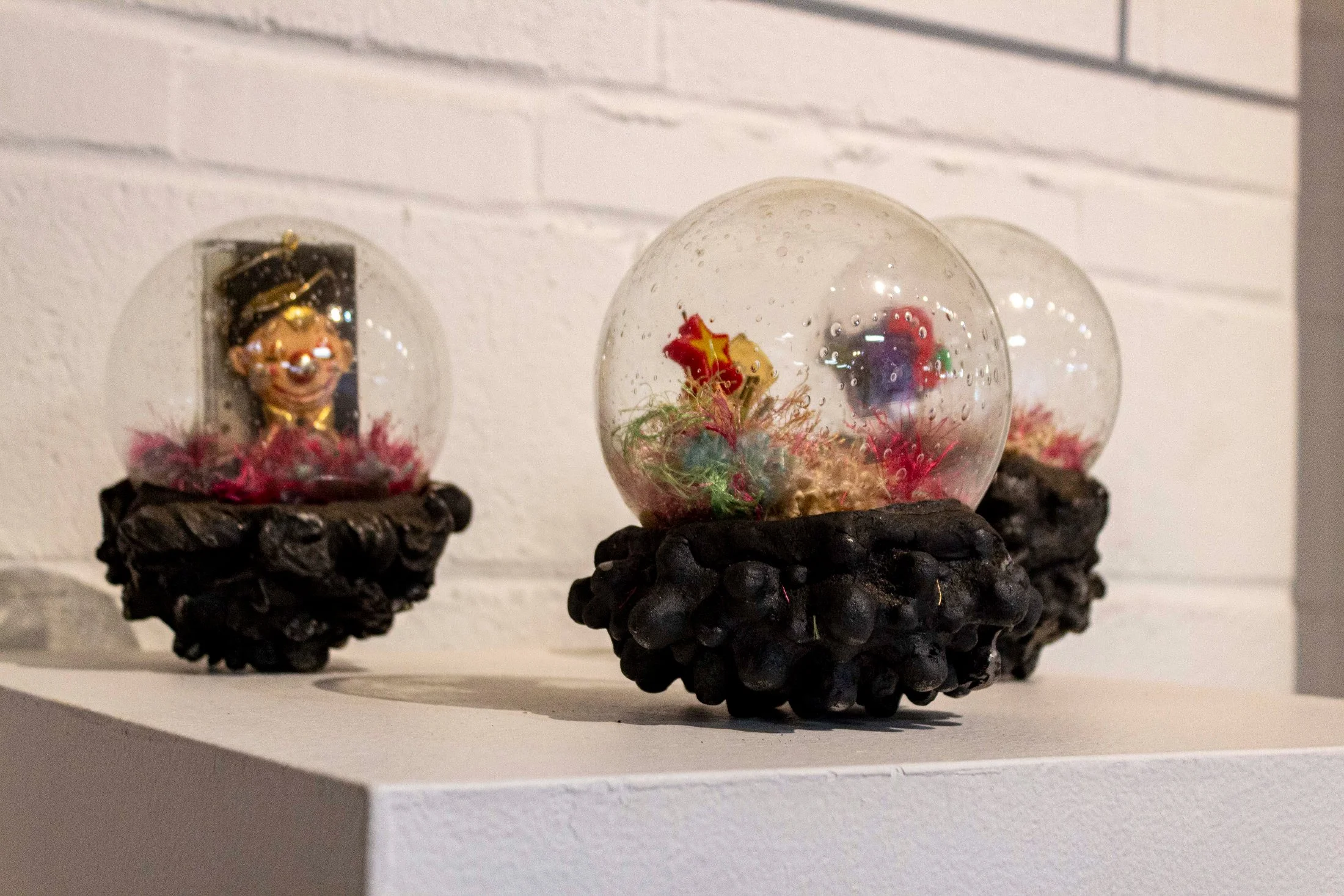 Three decorative snow globes on a white shelf, each featuring colorful and whimsical scenes inside, set against a plain white wall.