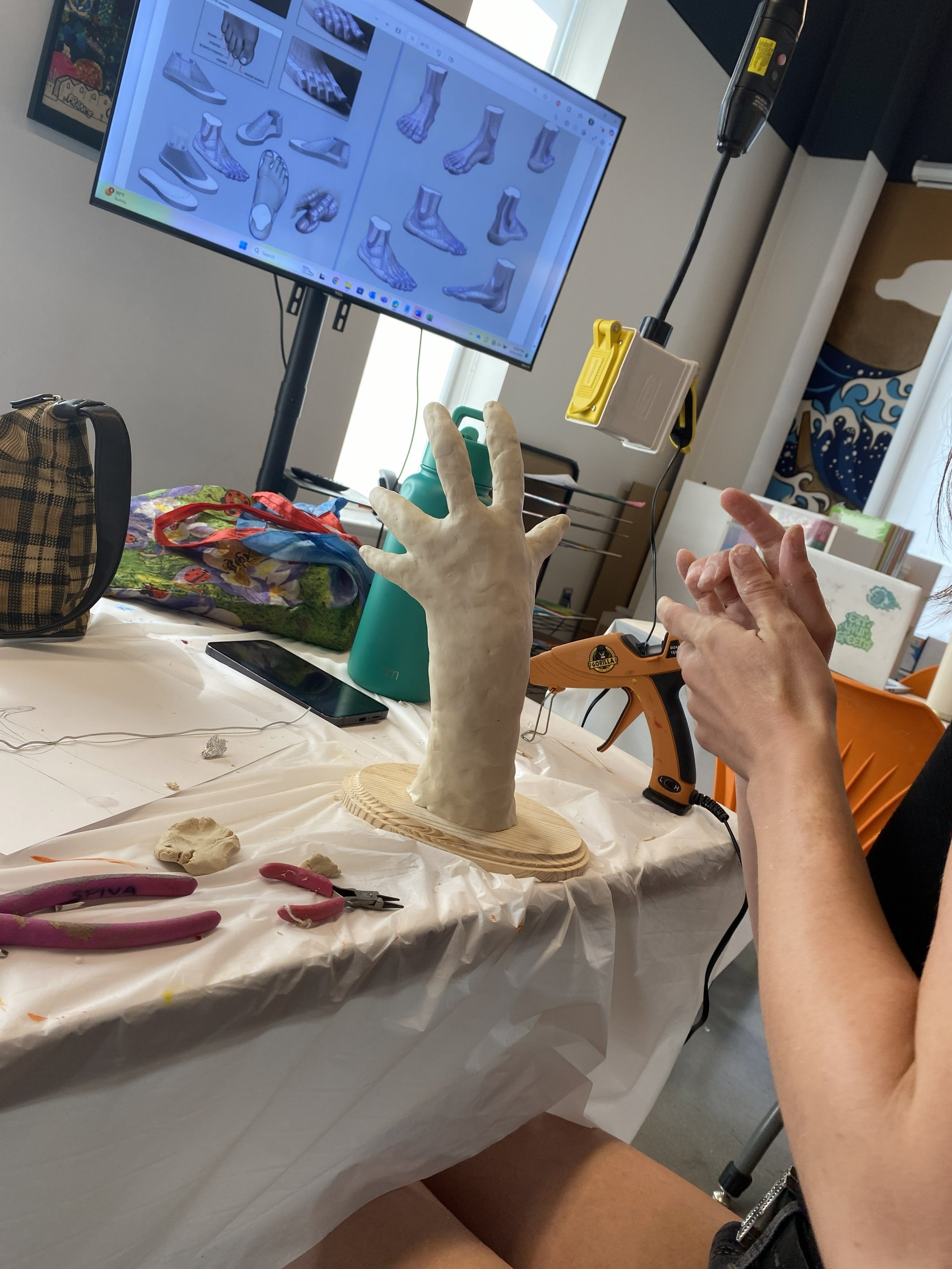 A sculptor's workspace with a mold of a hand, a hot glue gun, pliers, and clay on a work table, with a computer monitor displaying sketches of shoes and feet.