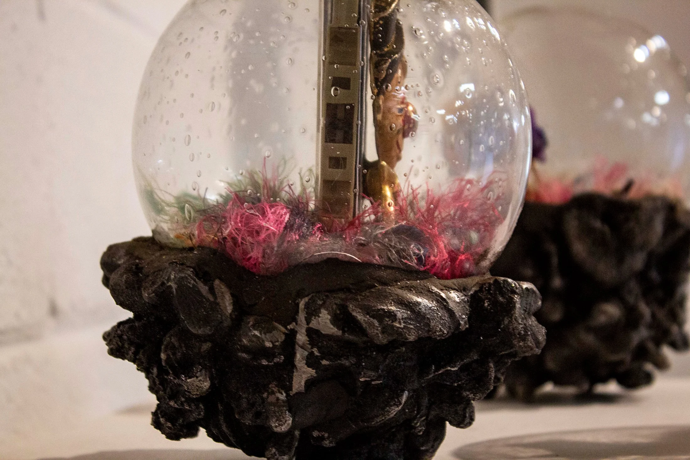 A glass orb with water droplets rests on a textured, dark base. Inside the orb, there are colorful fibers and a shiny metallic object, with a reflection of a person taking the photo.