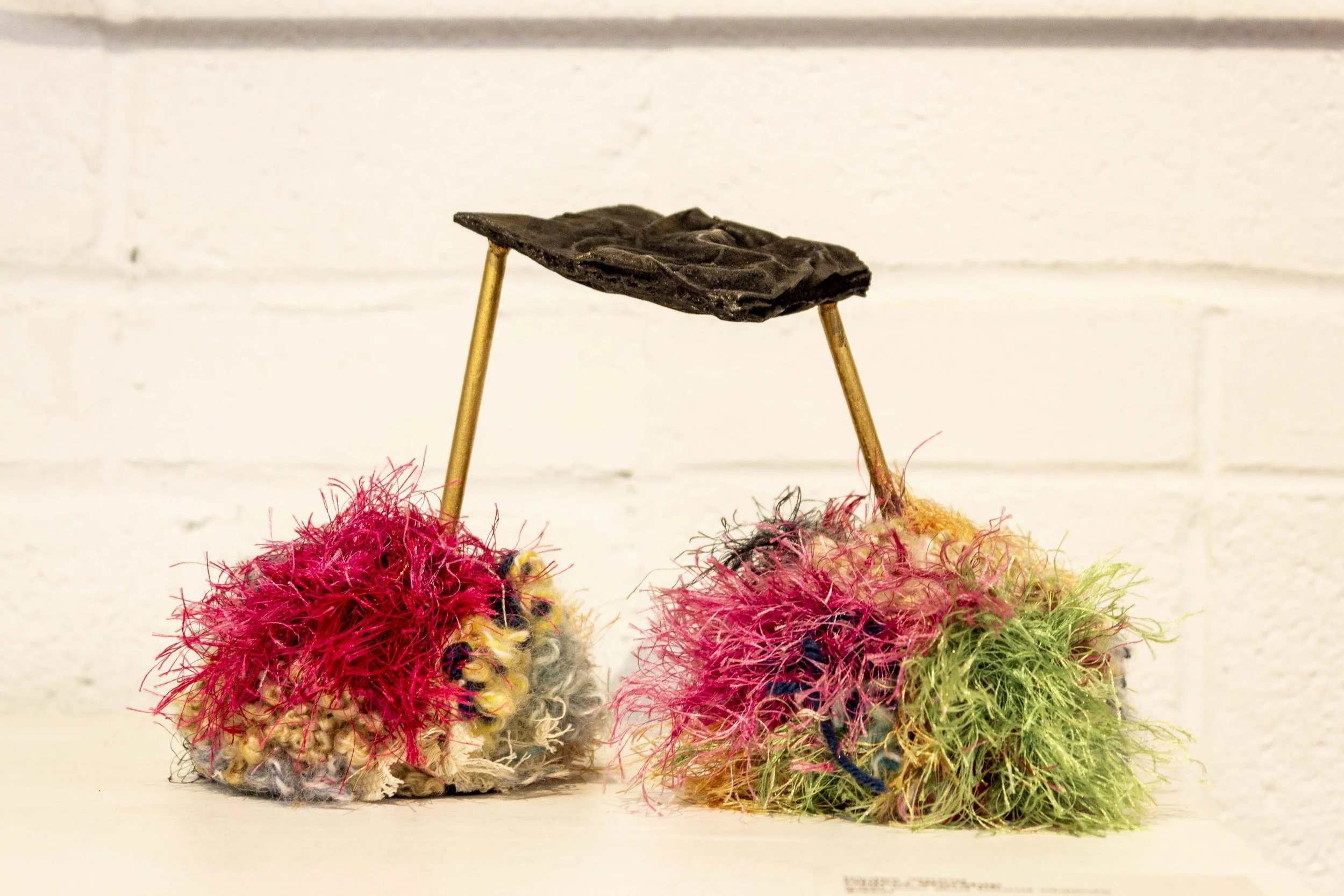 Close-up of colorful, fuzzy, pom-pom-like objects with a dark, textured object attached to two thin sticks above them, against a plain white background.