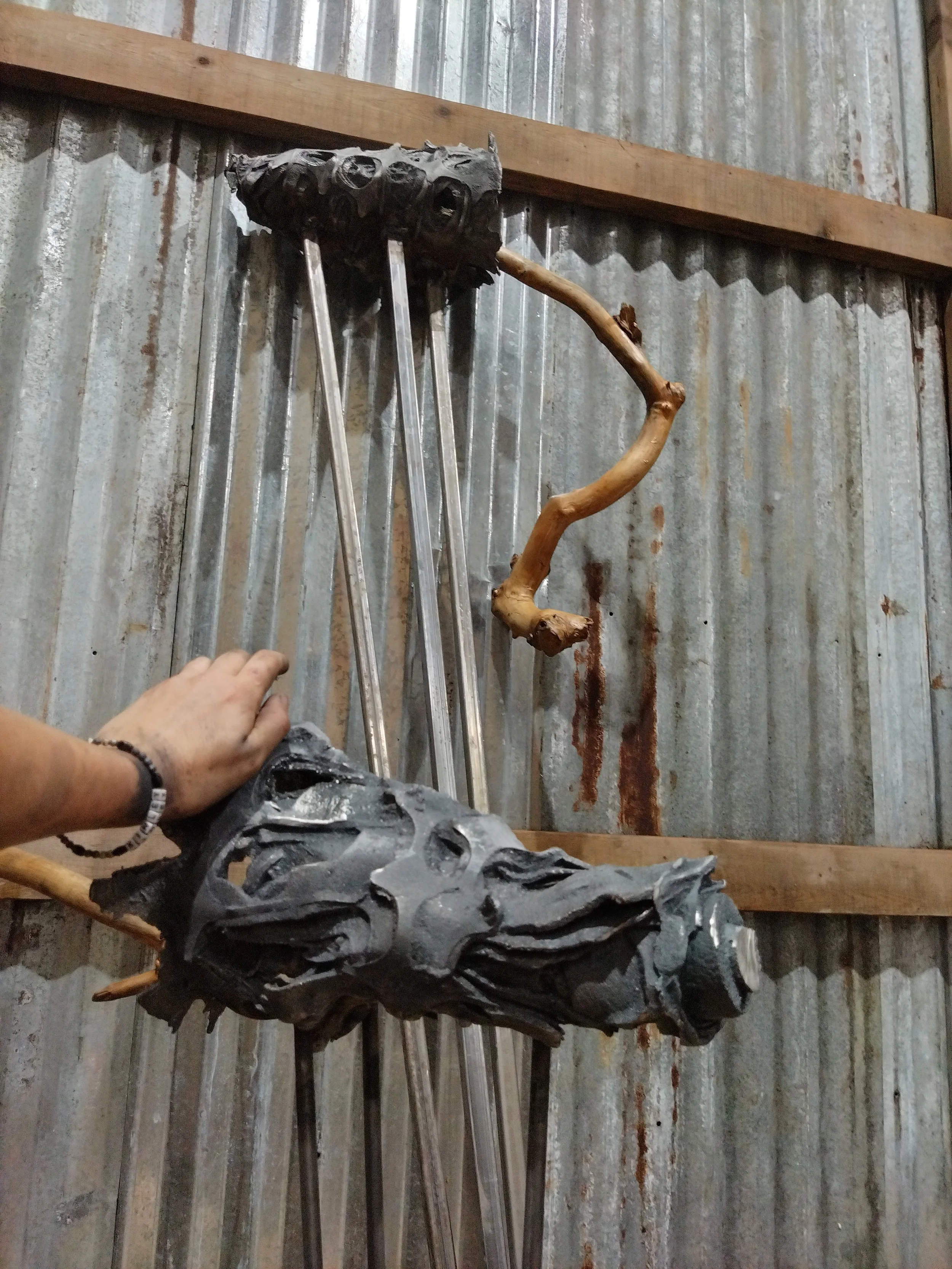 A person wearing a bracelet holding a black, textured object with multiple holes, with a wooden branch and metal tools leaning against a rusty corrugated metal wall.