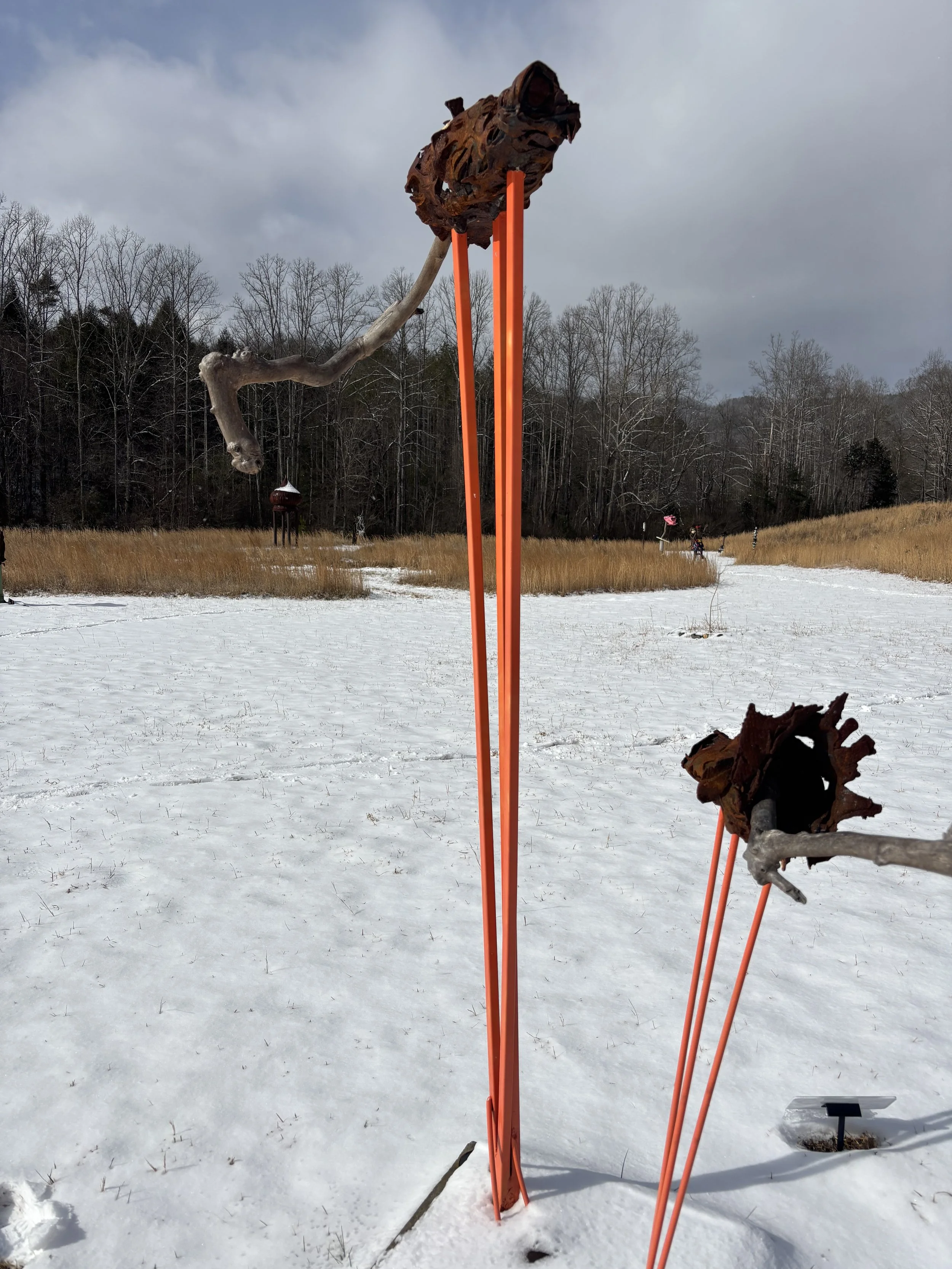 Outdoor art installation with a weathered metal sculpture of a cone of construction fencing supported by bright orange metal stands, set against a snowy landscape with trees and cloudy sky in the background.