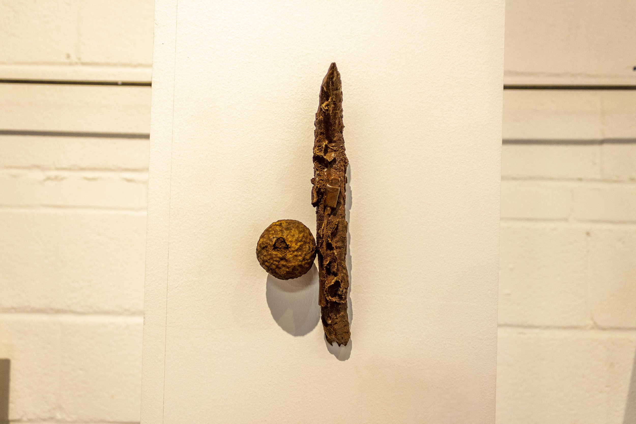 An art installation featuring a rusted, elongated metal object resembling a spear or tool, with a rusted, spherical object attached near the bottom, displayed against a white wall with horizontal lines.