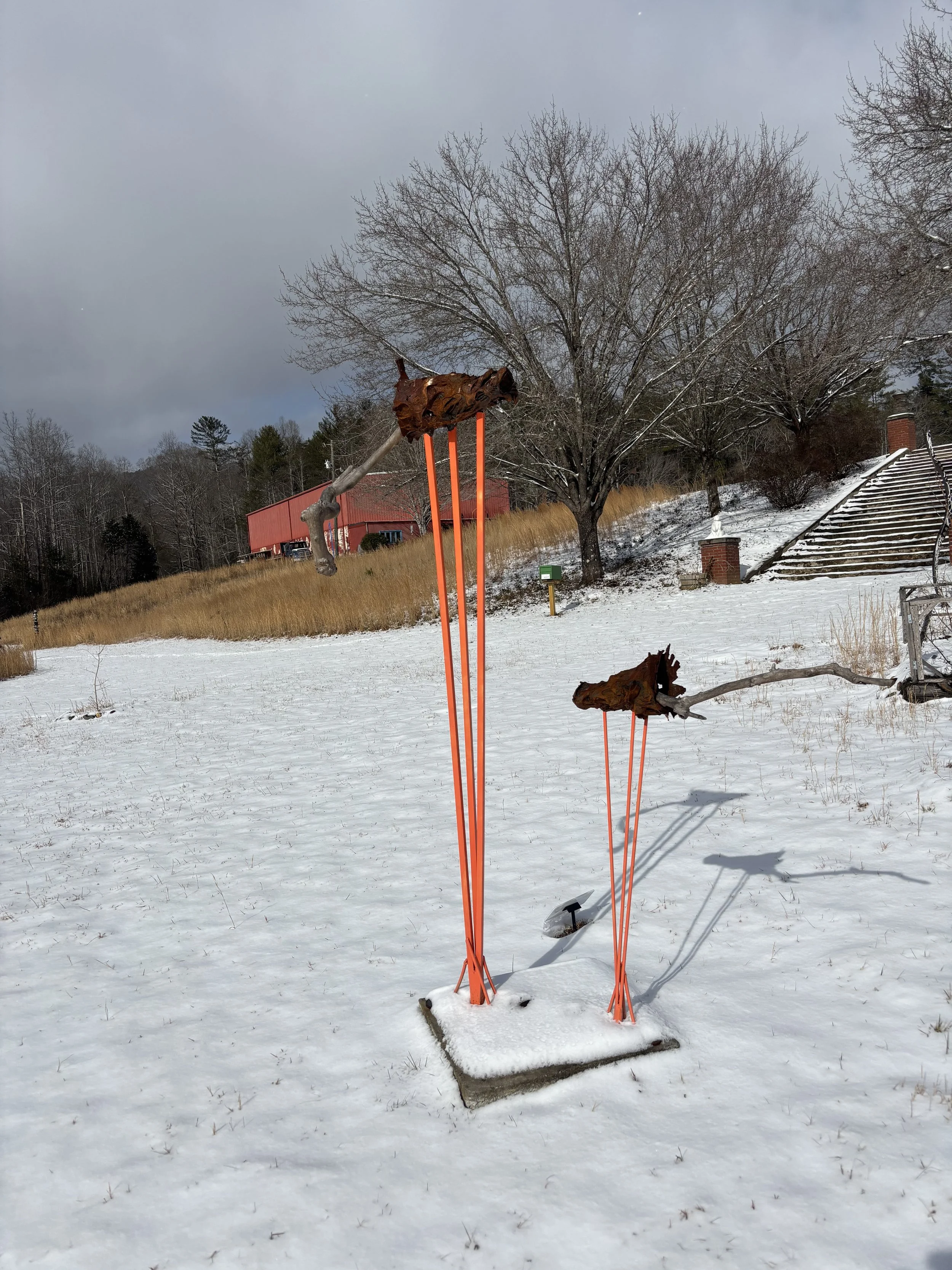 Outdoor scene with snow-covered ground, leafless trees, a staircase, a red building, and a rusty sculpture resembling a dog on orange poles.