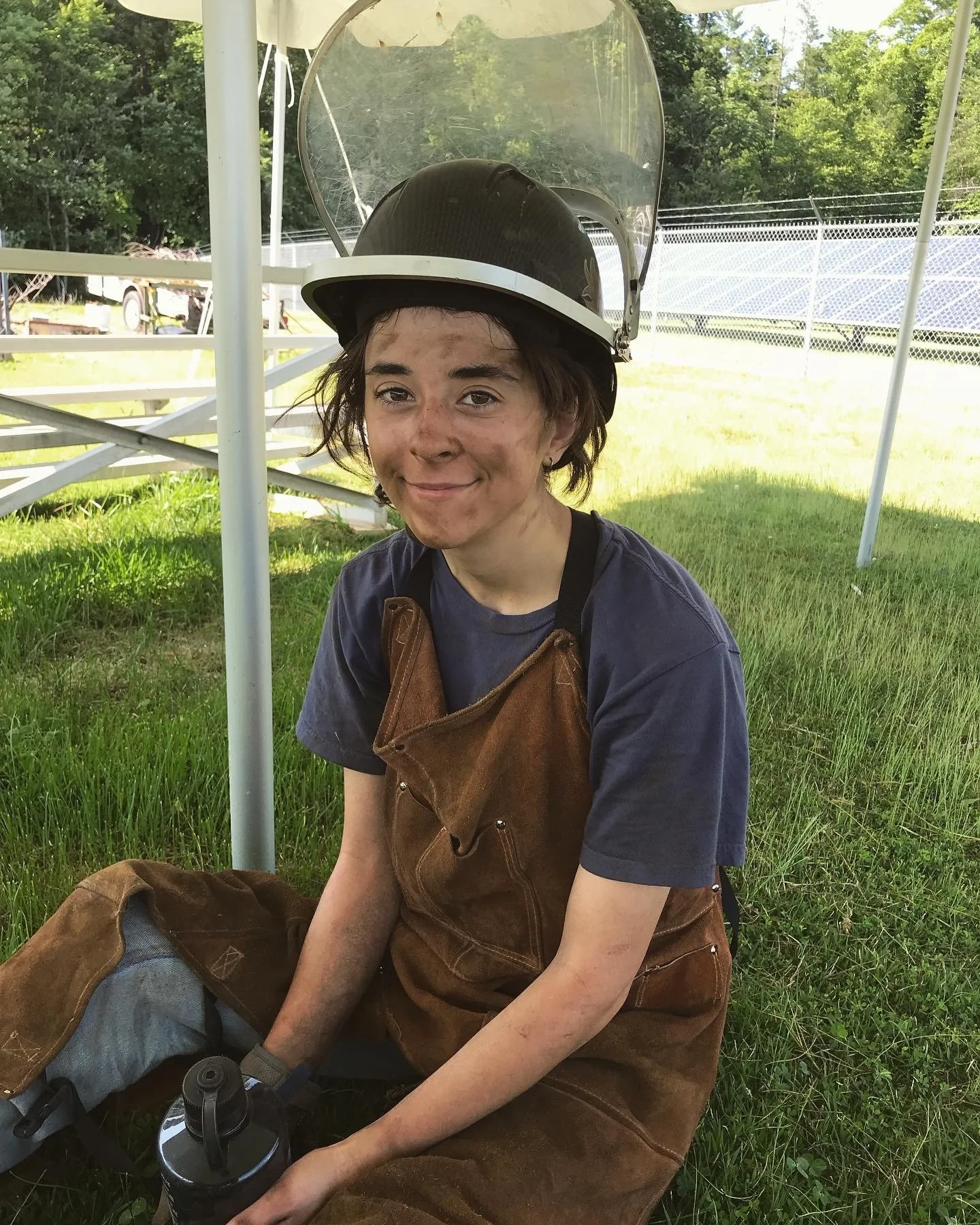 A young person with short hair sitting on grass, wearing a black helmet with a clear face shield, a brown apron, and a navy blue t-shirt, smiling at the camera. Background shows a fenced outdoor area with solar panels and trees.