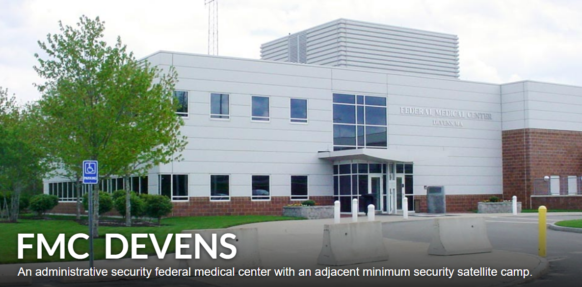 Front view of a federal medical center building with a sign that reads 'Federal Medical Center.' There are trees, bushes, and a handicap parking sign in the foreground, with concrete barriers on the road leading to the entrance. SOMP Yard.