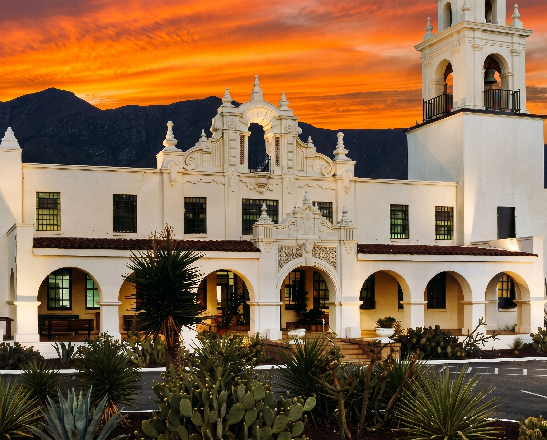 A white Spanish-style building with a bell tower, arches, and decorative architectural details, set against a vibrant orange sunset sky with mountains in the background. In the foreground, there are various desert plants including cacti and succulents.