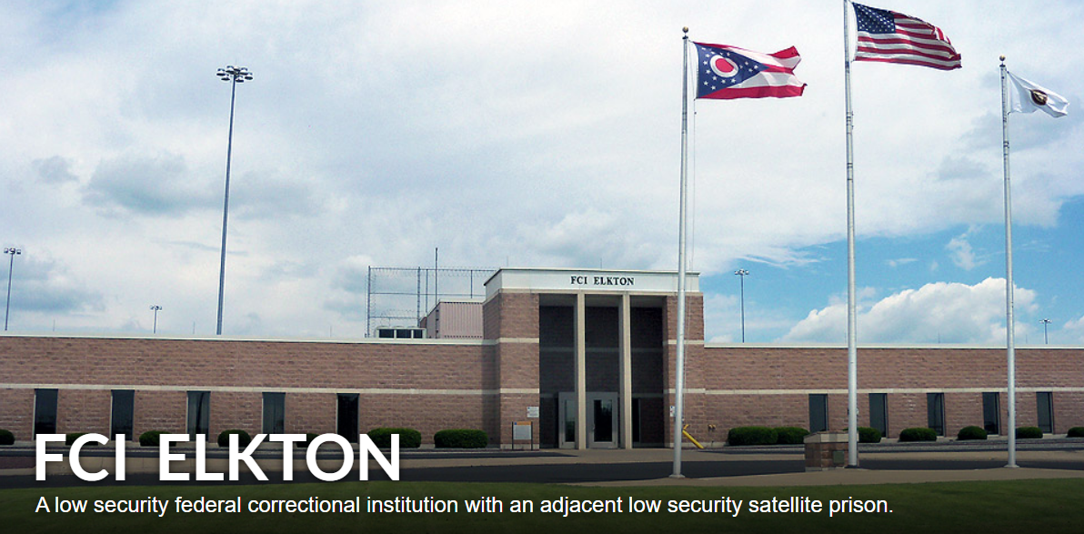 Front view of FCI Elkton low security federal correctional facility with three flags flying, surrounded by a parking lot and a grassy area, under a partly cloudy sky. SOMP Yard.