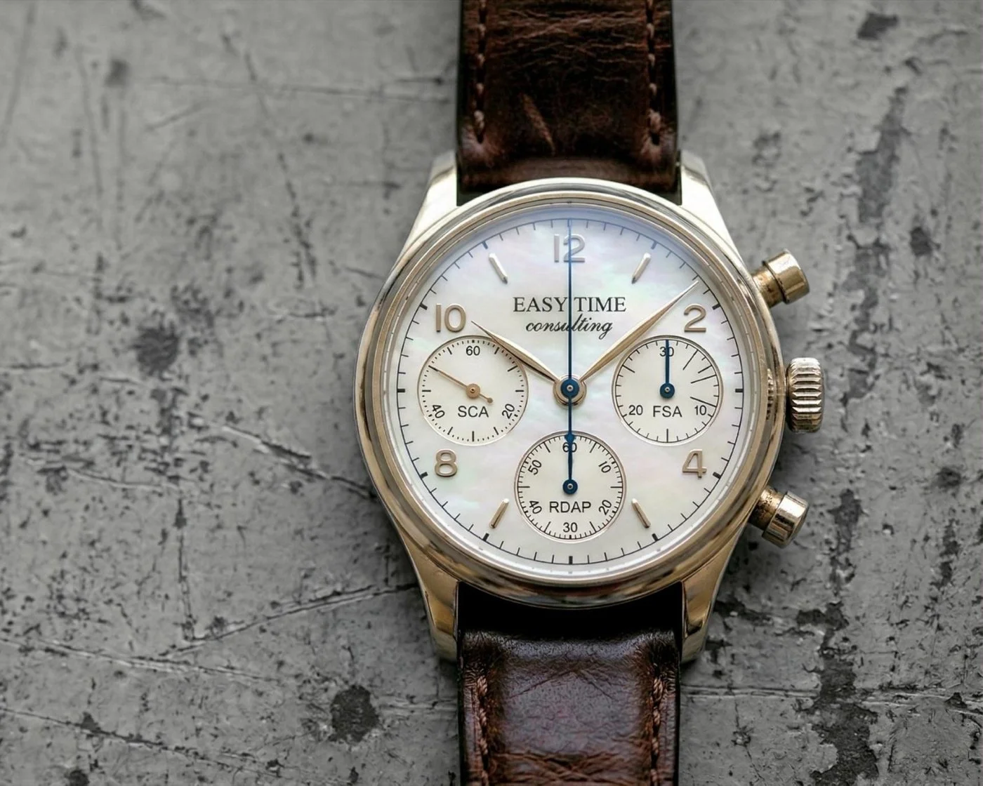 High end chronograph watch displayed head-on from a distressed metal sheet. The dial face has signature text that reads Easy Time Consulting, There are 3 sub-dials with the signatures SCA, RDAP, and GTC.