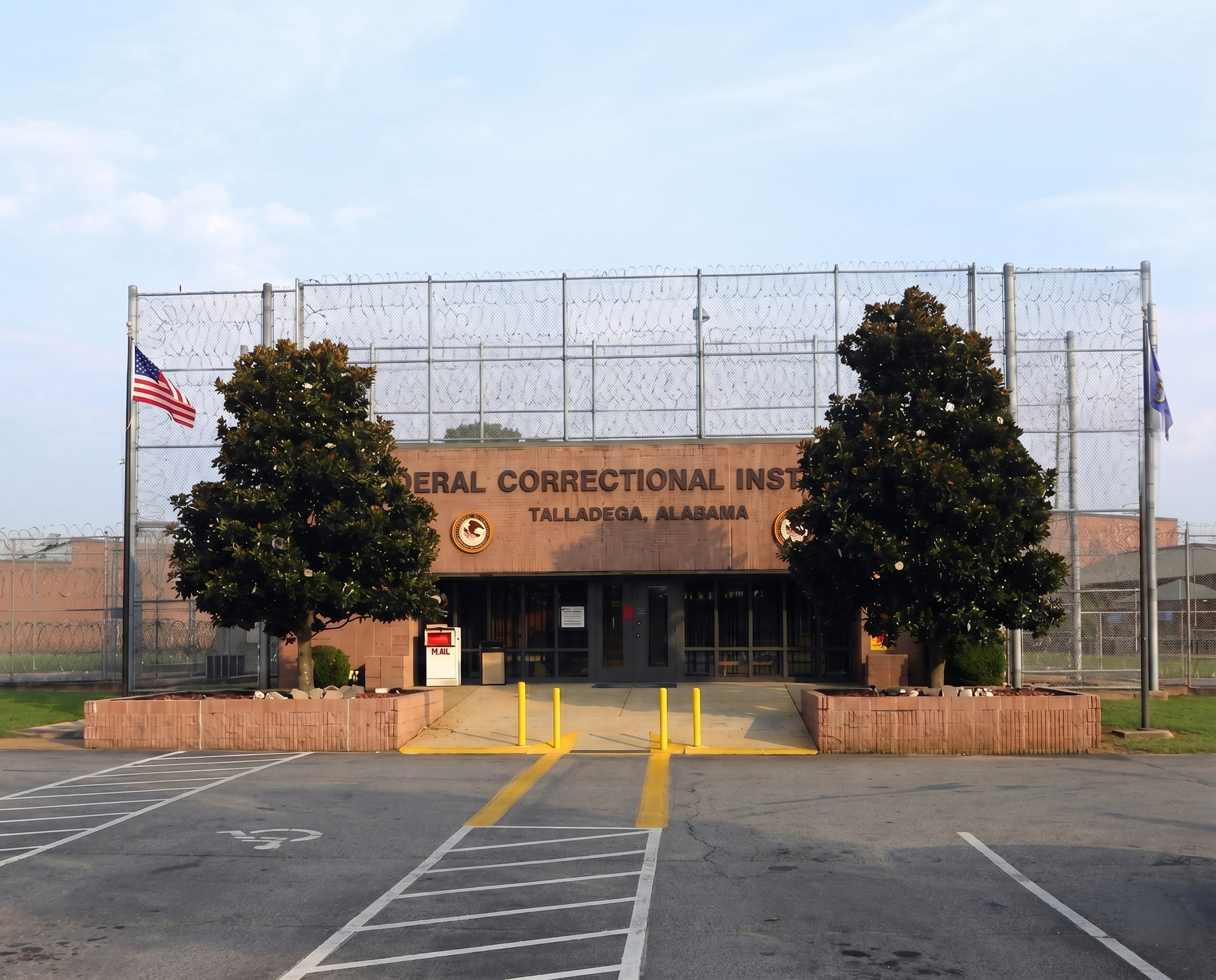 The entrance of the Federal Correctional Institution in Talladega, Alabama, featuring brick walls, a chain-link fence with barbed wire on top, and American flags on either side. There are trees in front, parking space with painted lines, and a sign above the doorway.
