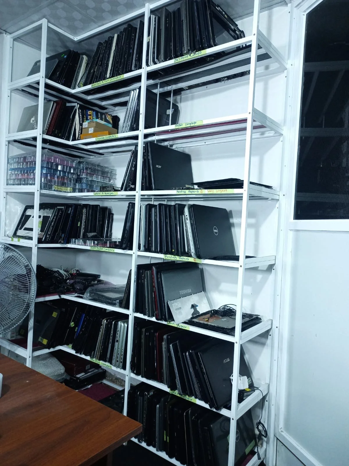 White metal shelving unit filled with organized laptops, electronics, and storage boxes, with labels indicating repair and computer project areas. A fan is visible on the left and a wooden table in the foreground.