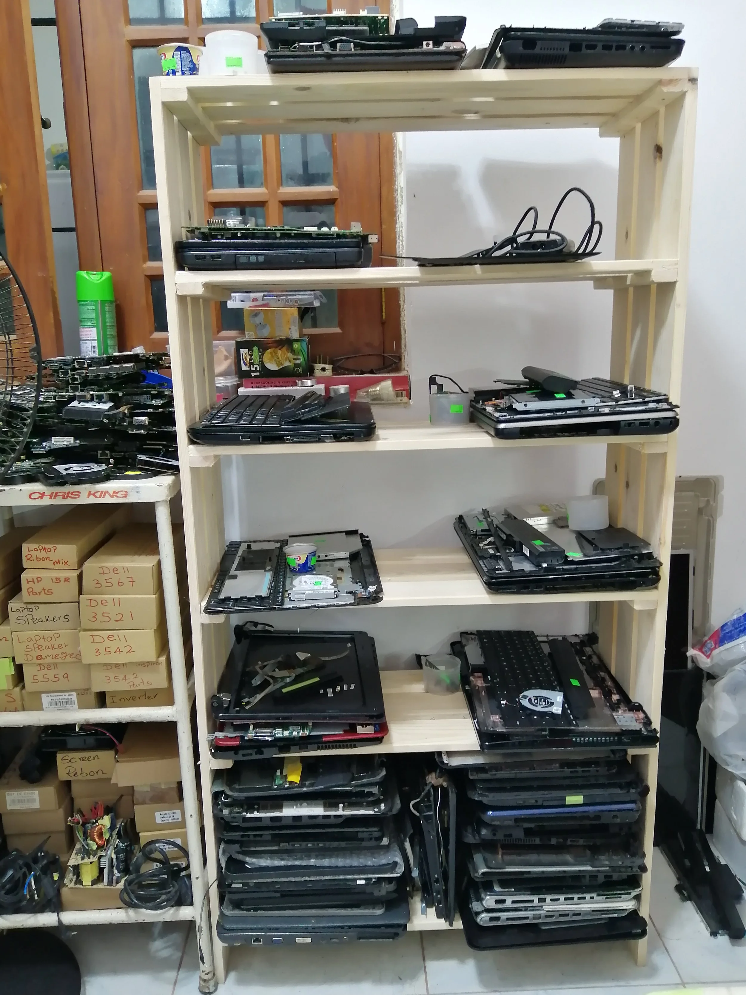 Wooden shelving unit filled with disassembled laptops, computer parts, and electronics. Boxes with labels on the side are stacked to the left.
