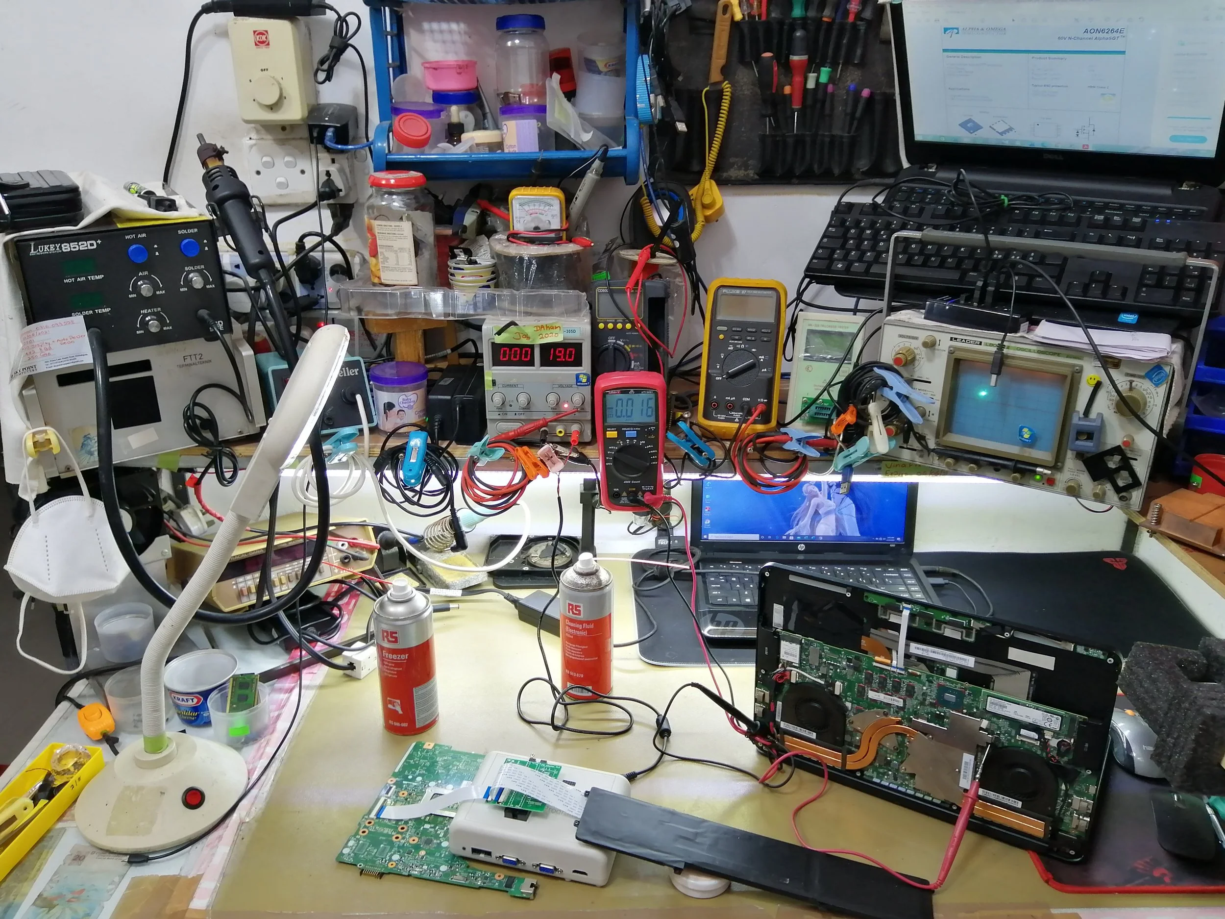 A cluttered workbench with electronic tools, circuit boards, and computers, surrounded by various small containers, tools, and testing equipment.