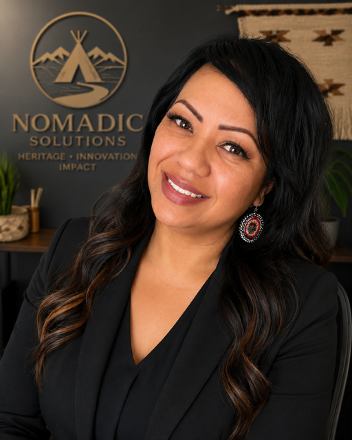 Professional woman with dark, wavy hair wearing a black blazer and red earrings, smiling in an office with a logo that says 'Nomadic Solutions' in the background.