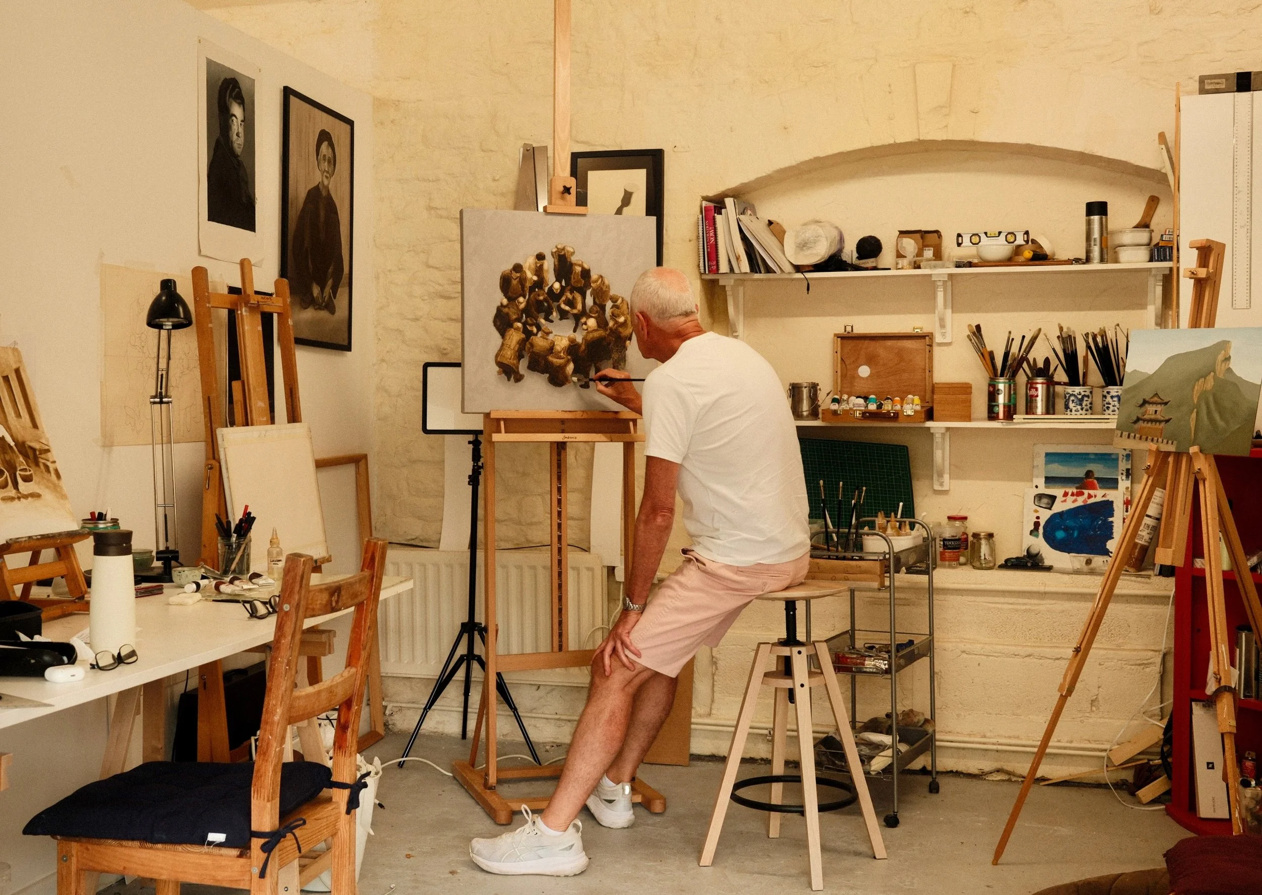 An older man with white hair in a white t-shirt and pink shorts is painting an art piece in a studio. The studio has various artworks, shelves with supplies, and easels.