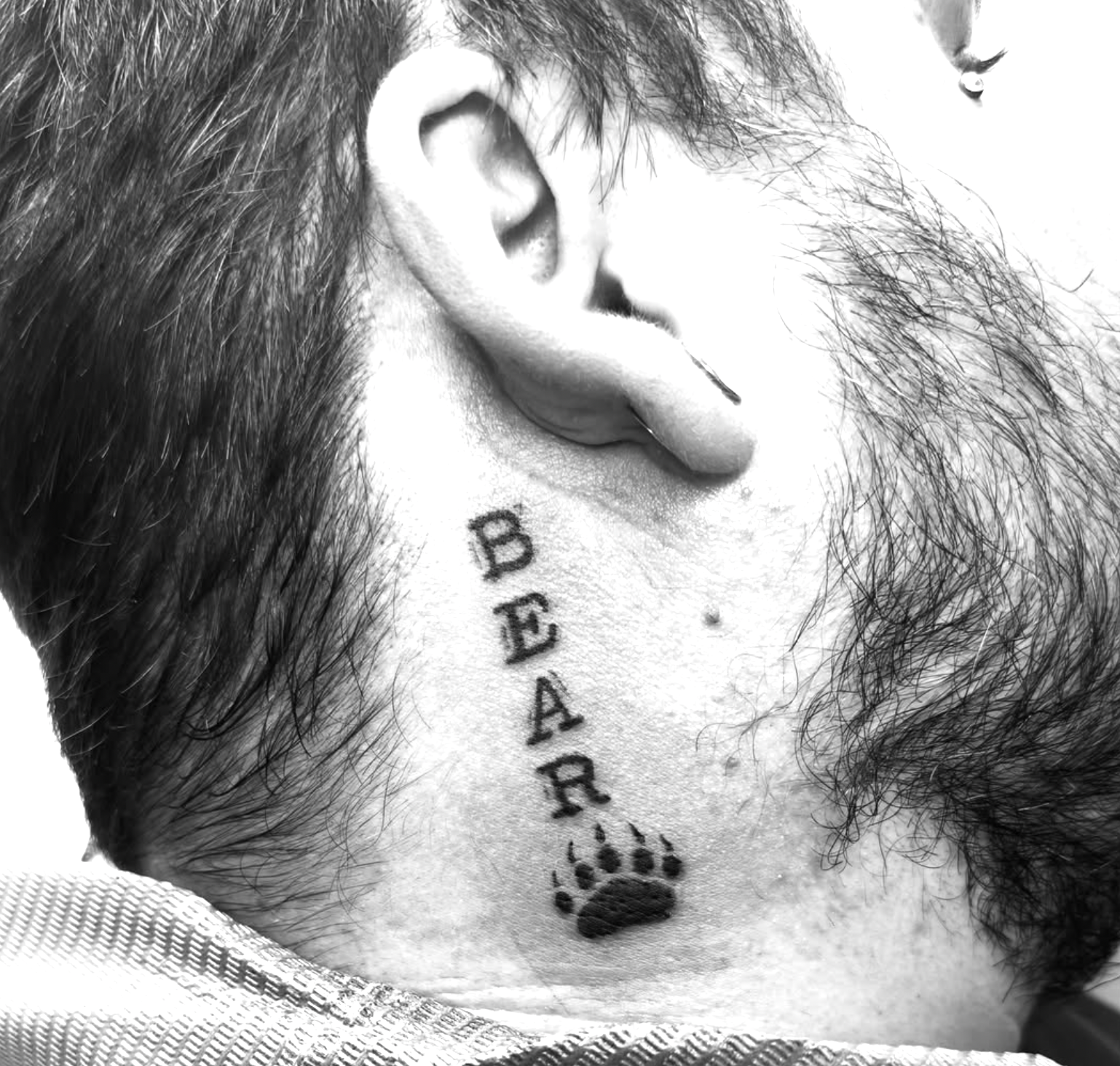 Close-up black and white photo of a person's neck and lower face, with tattoos reading 'BEAR' and a paw print, ear with an earring, facial hair, and part of their hair visible.