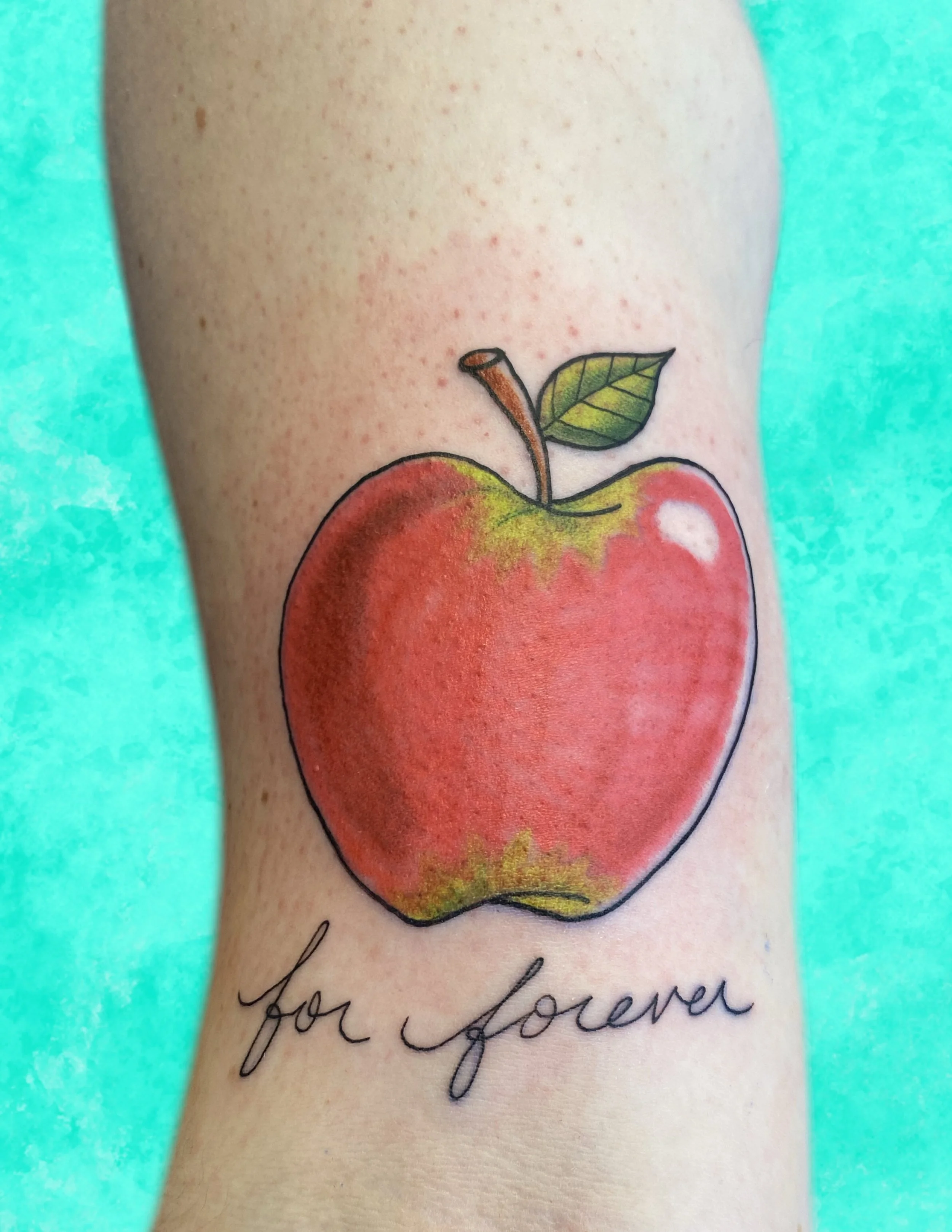A tattoo of a red apple with a green leaf and a brown stem on a person's arm, along with a handwritten message that partially reads 'for j...'