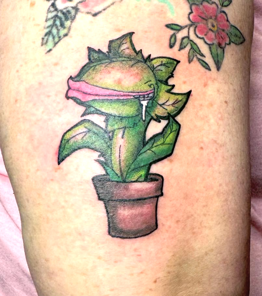 Tattoo of a plant with a carnivorous, Venus flytrap-like appearance in a flowerpot, with a smaller flower tattoo above it.