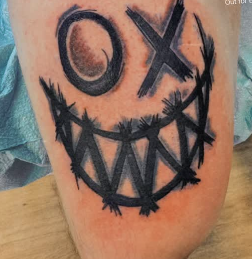 Tattoo of a smiley face with a crescent moon for a eye and the other eye in the shape of an X, with a wide, jagged smile.