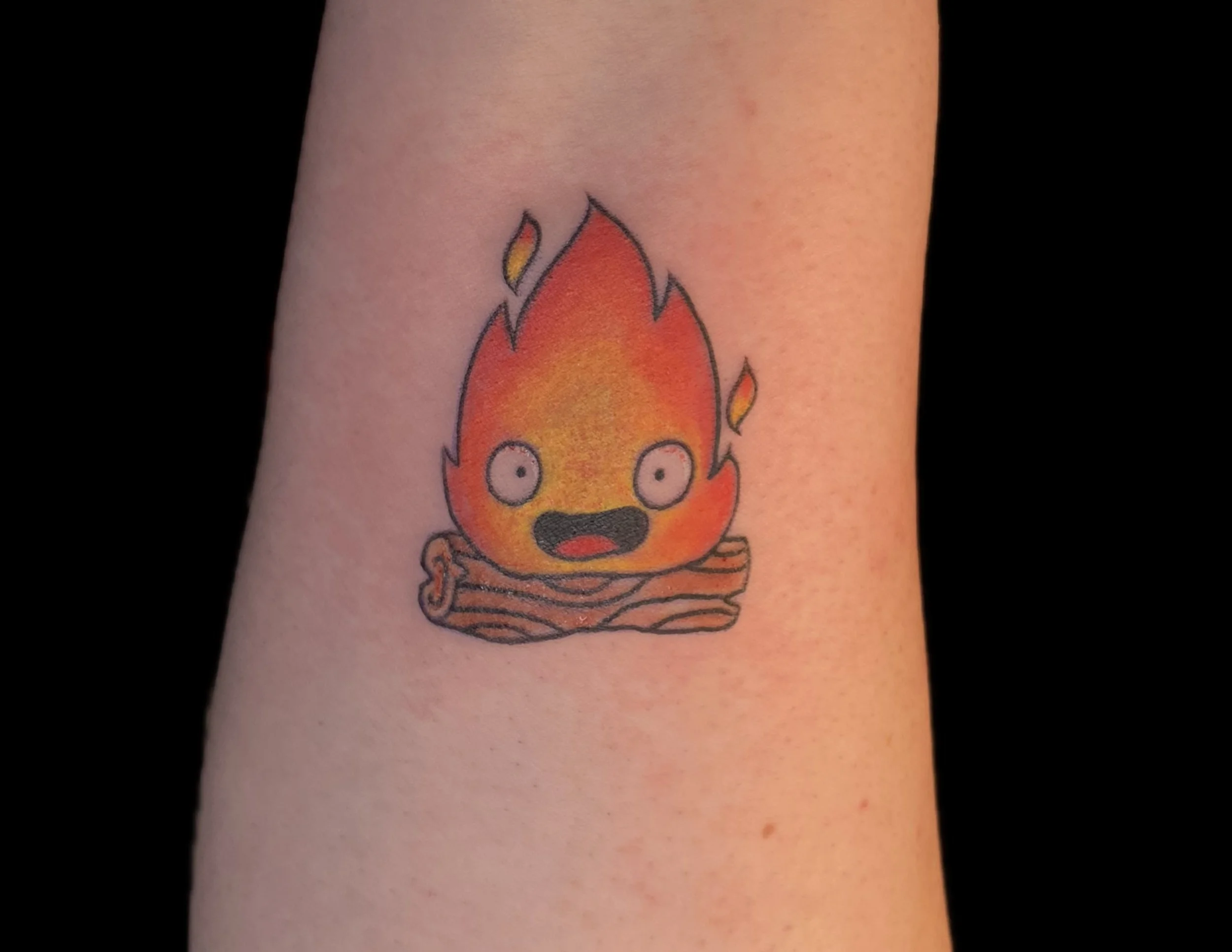 Tattoo of a cartoon flame with eyes, mouth, and a striped base on person's skin.