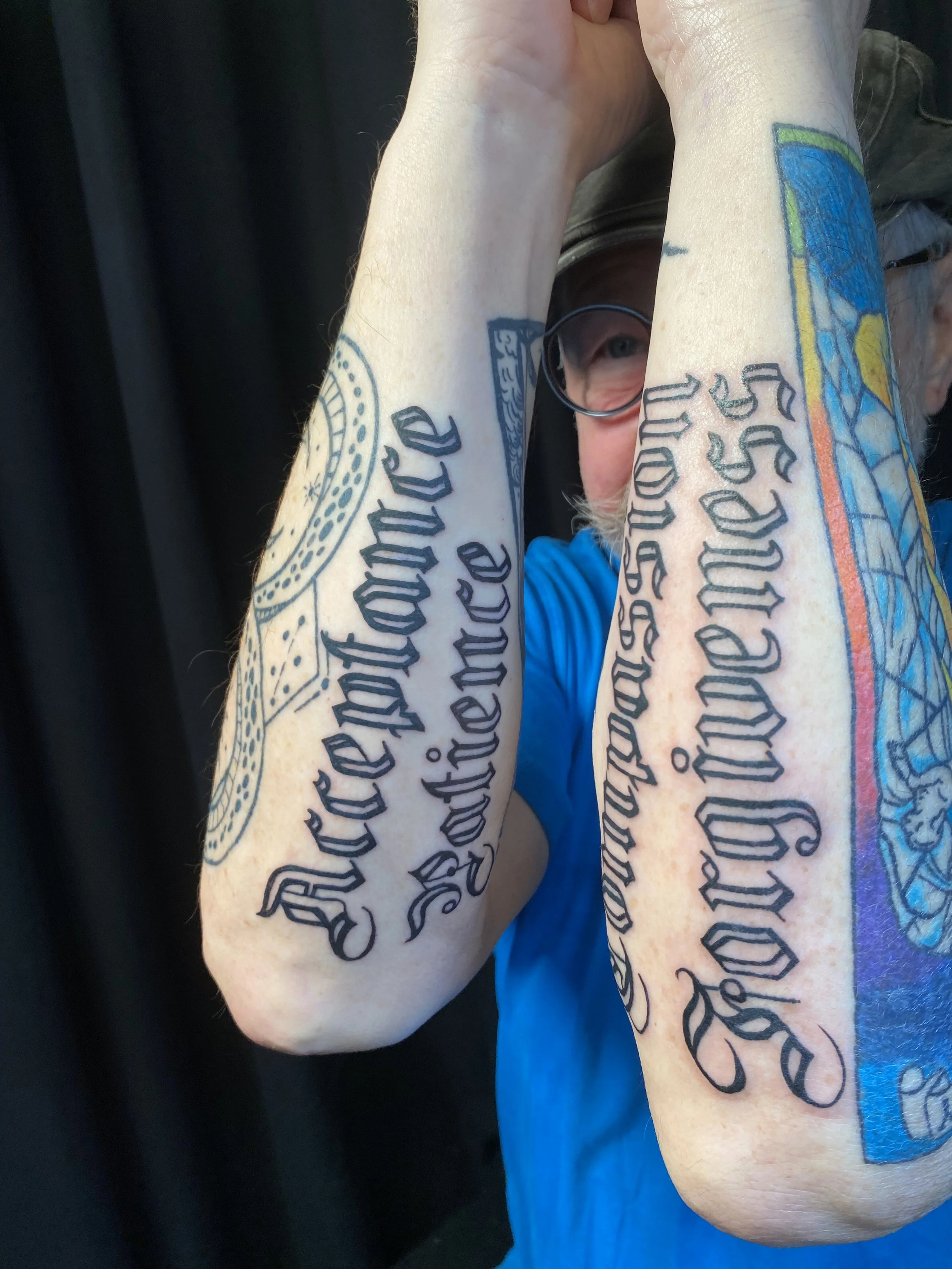 Close-up of a person's arms covered in colorful tattoos with black script text, appearing to be a person with glasses and a blue shirt.