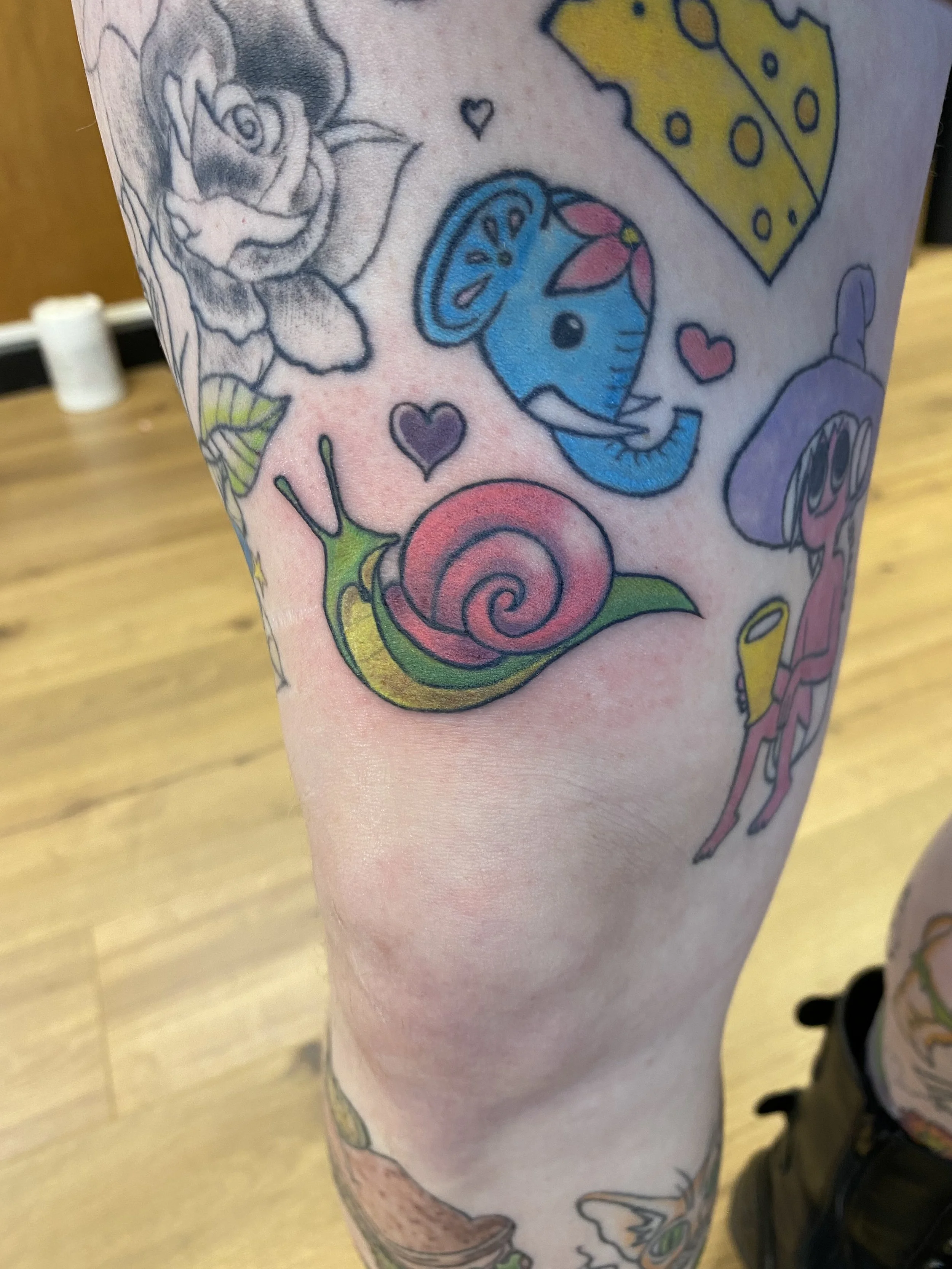 Colorful tattoos on a person's arm featuring a rose, a slice of cheese, a blue horse, hearts, a snail, and other whimsical designs.