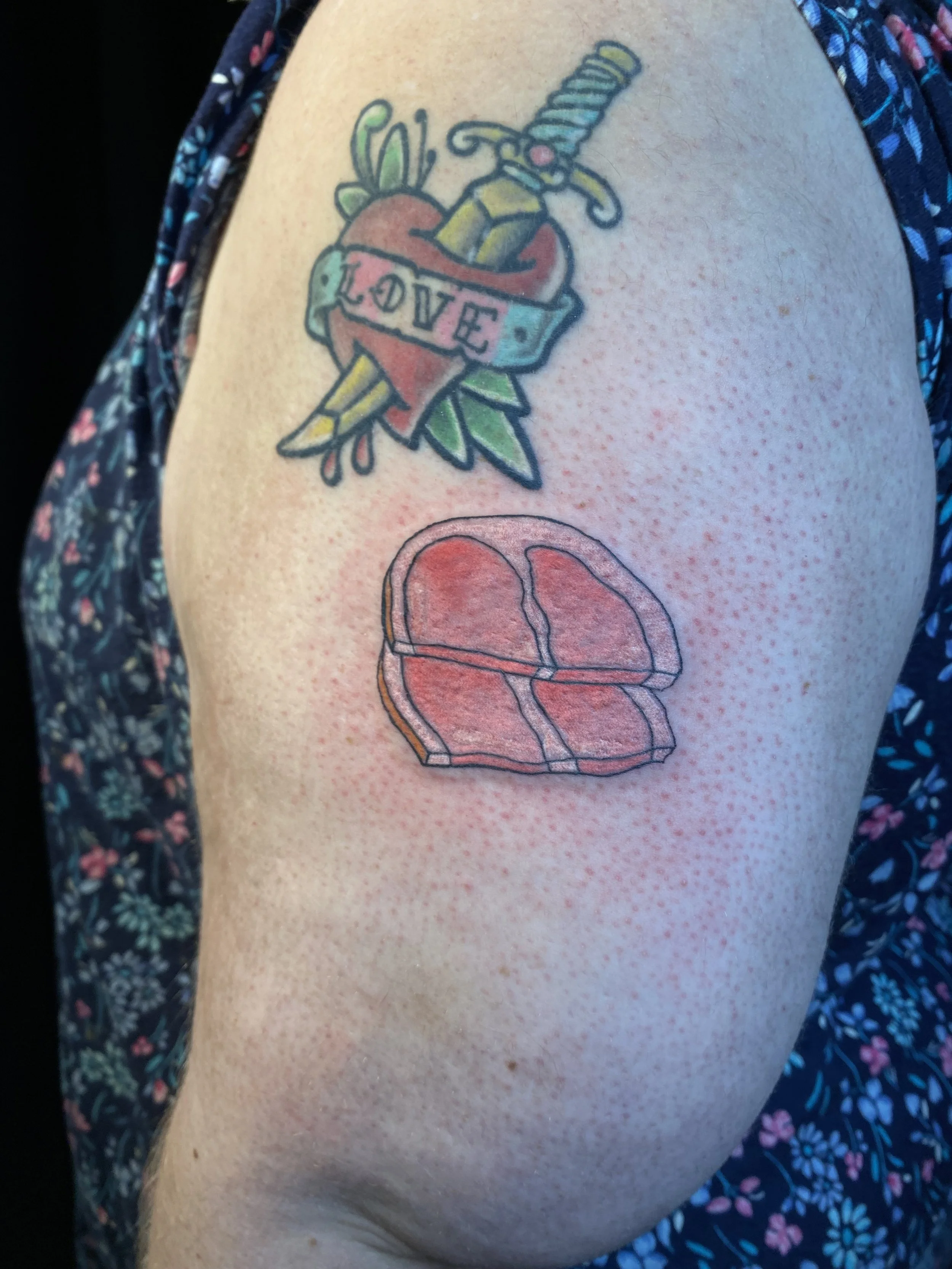 Colorful tattoo of a heart with a banner that says "LOVE," an arrow piercing through it, and a sword behind the heart, on a person's upper arm.