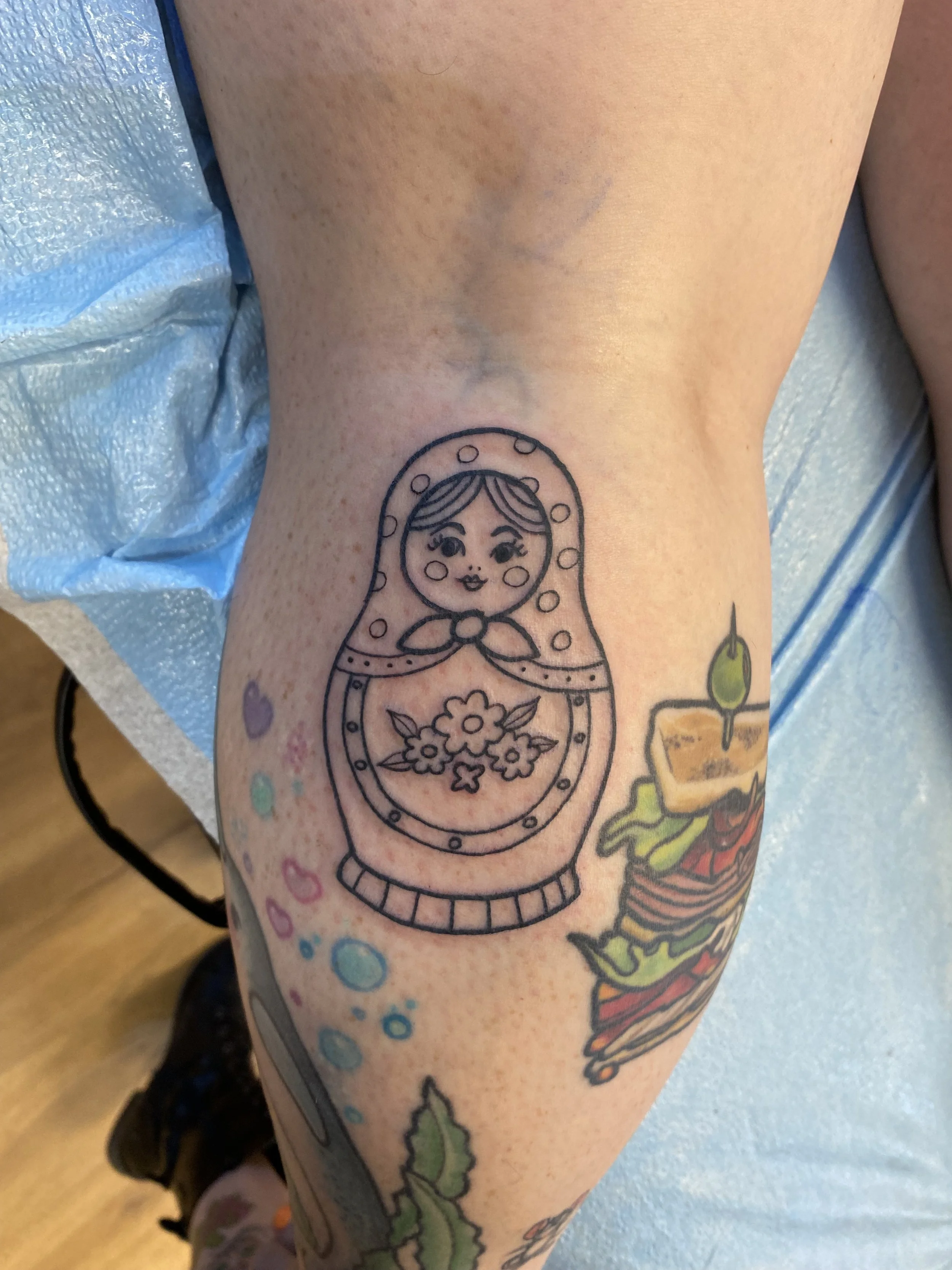 A tattoo of a matryoshka doll with flowers on a person's calf, surrounded by smaller colorful heart and circle designs.