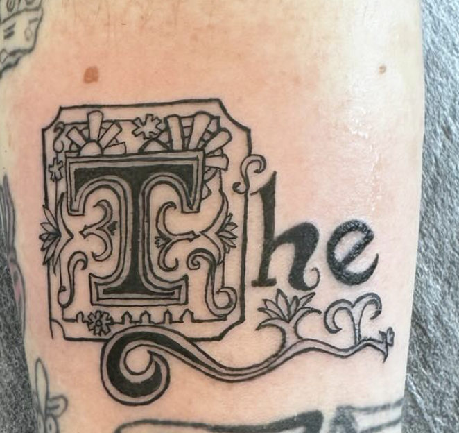 Close-up of a tattoo on skin, featuring an ornate capital letter 'T' with decorative flourishes, and the word 'he' in cursive.
