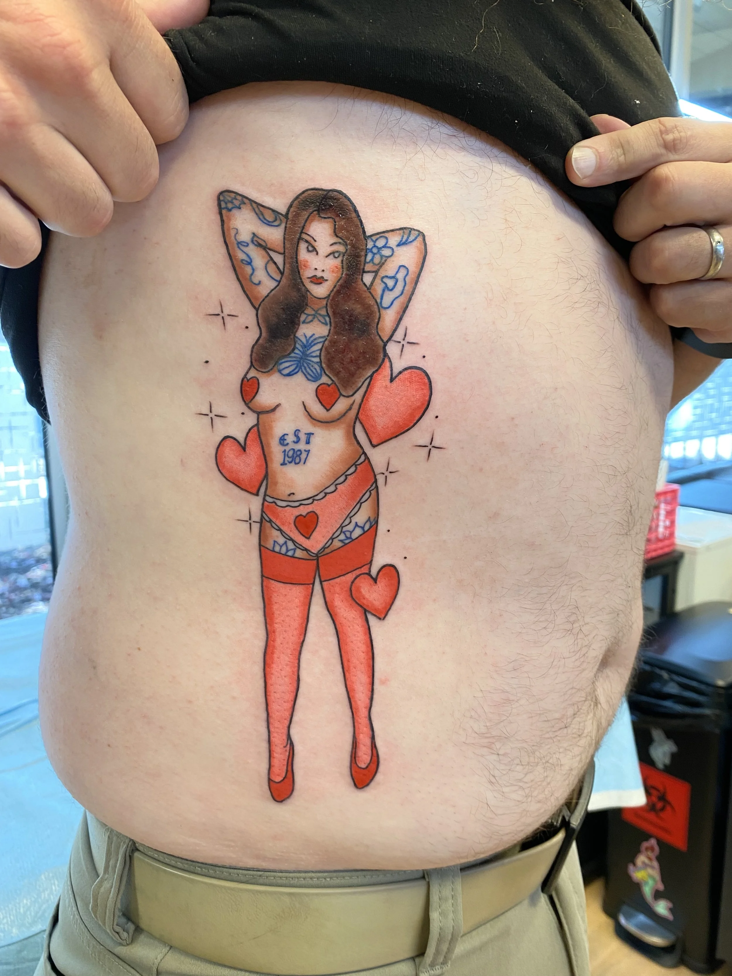 Tattoo of a woman with long hair in lingerie surrounded by hearts and stars on person's side.