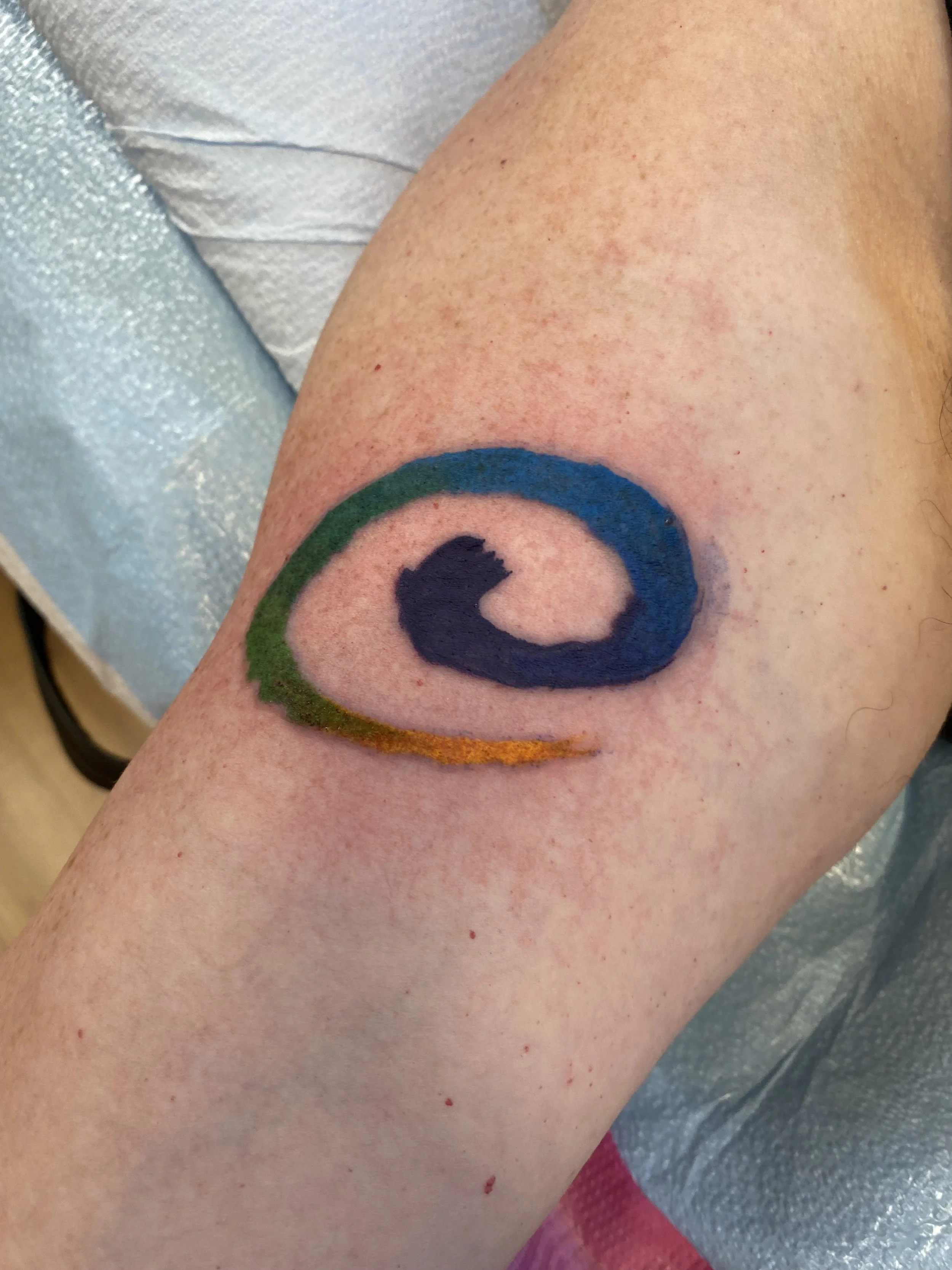 A person's arm with a colorful ink tattoo of a target symbol, featuring blue, green, orange, and black colors, resting on a medical table or surface.