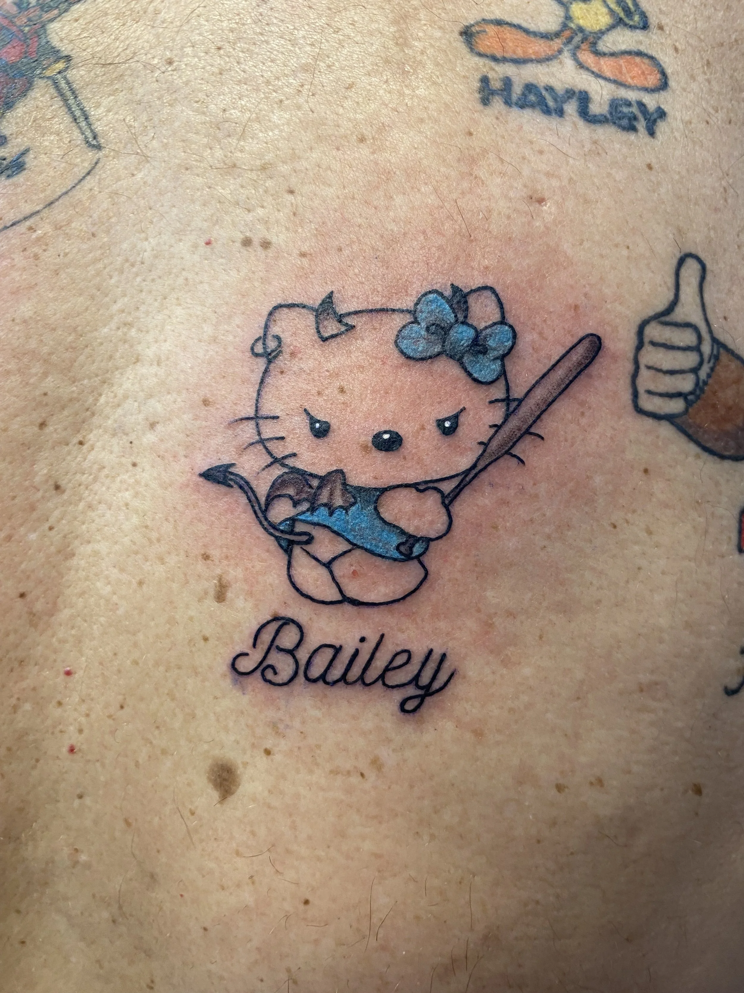 A close-up of a tattoo featuring Hello Kitty holding a spoon with a blue bow on its head, and the name "Bailey" written below.