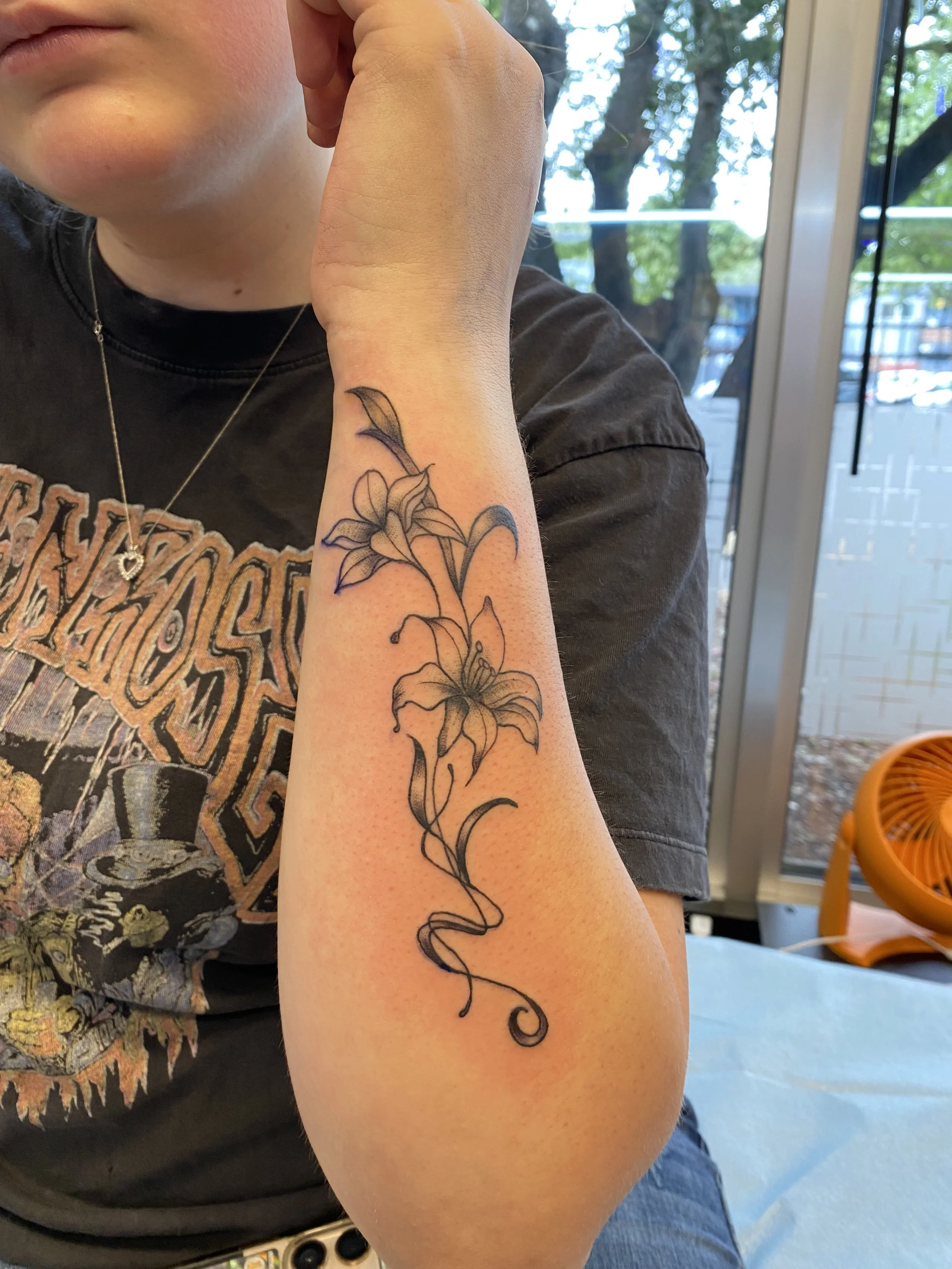 A person's arm with a tattoo of a lily flower and a trailing vine, sitting indoors near a glass door with trees visible outside.
