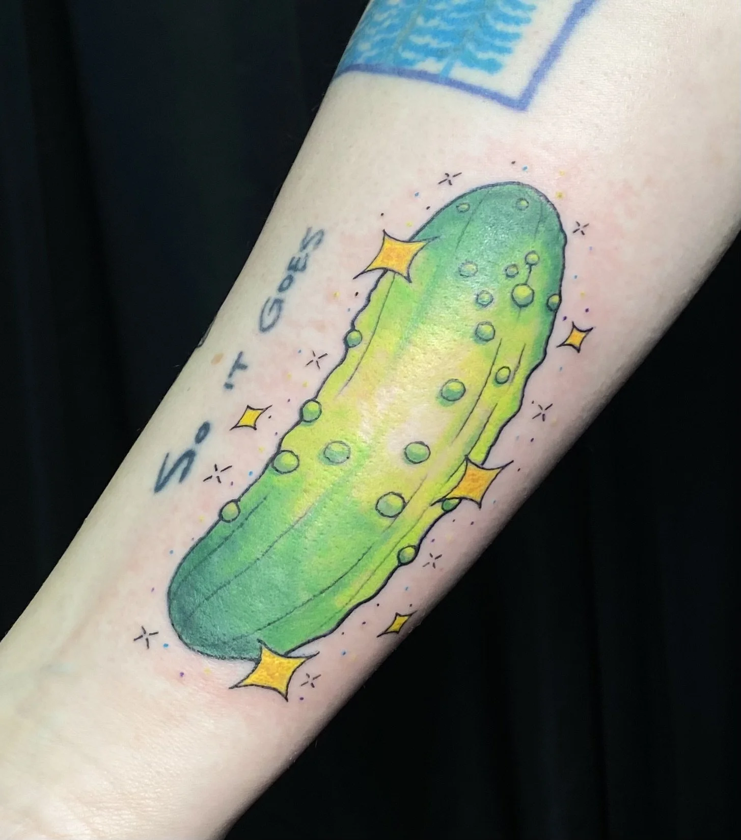 Colorful tattoo of a fluorescent green cucumber with yellow stars, tiny sparkling details, and the words 'SHIIT GEEEKS' written beside it.