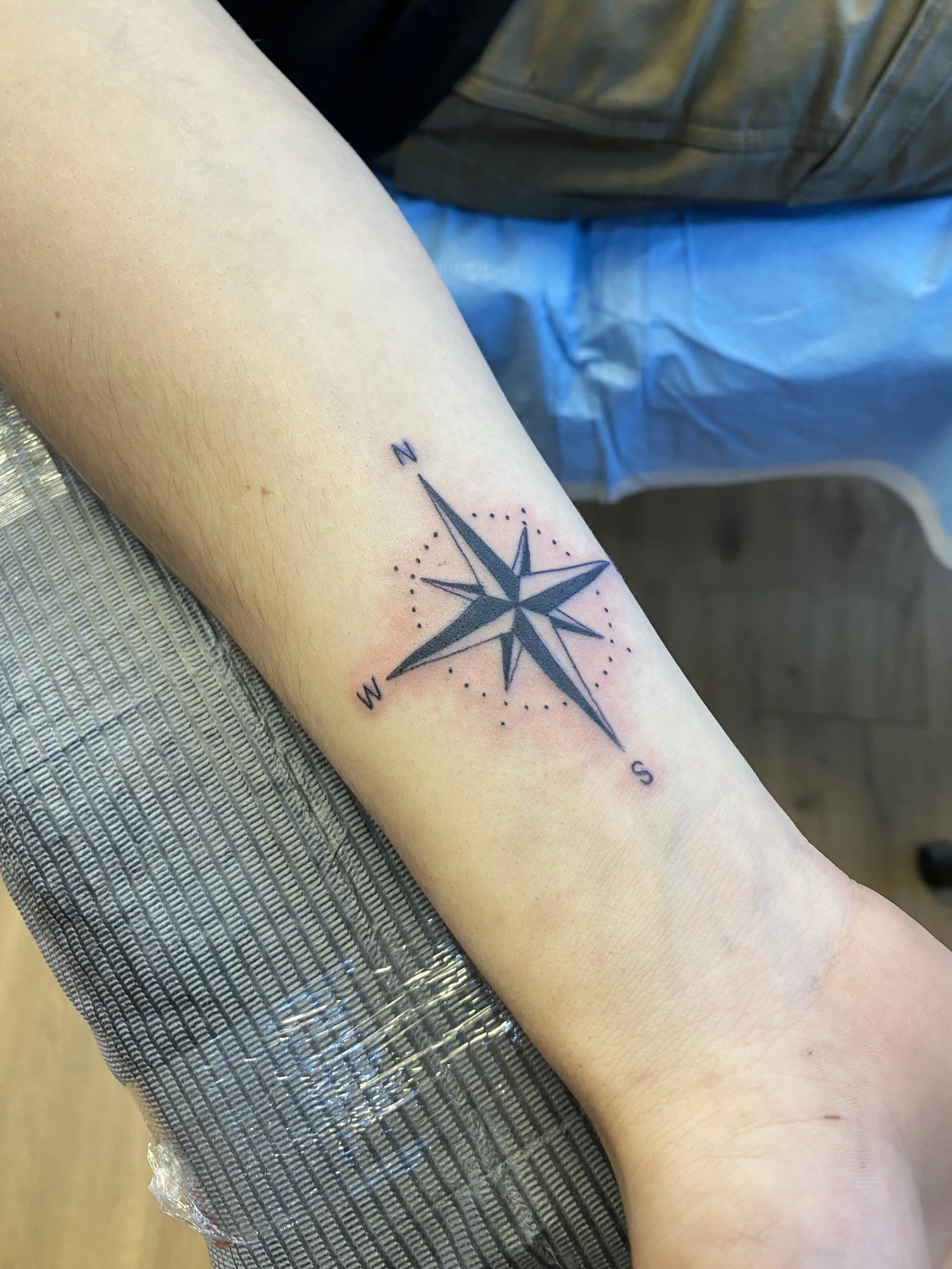 Close-up of a person's inner forearm with a freshly done black nautical compass tattoo surrounded by small dotted lines and labeled with 'N', 'S', 'E', and 'W' for directions.