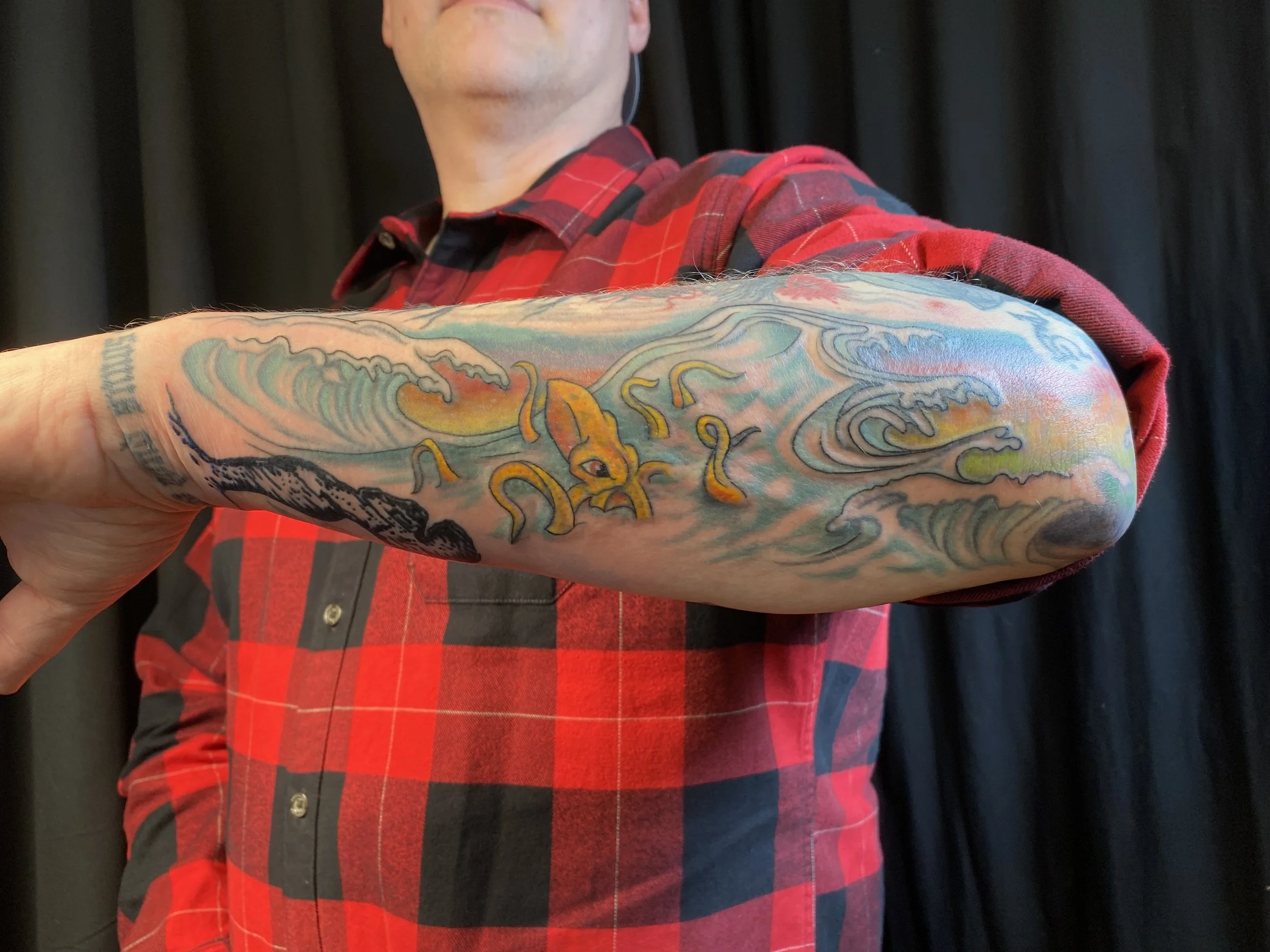 A man in a red plaid shirt showing his colorful tattooed arm in front of a black curtain.