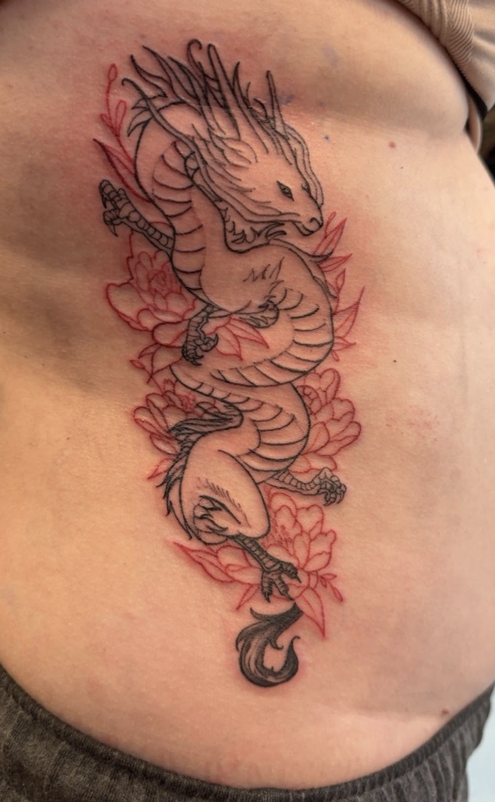 Tattoo of a dragon intertwined with flowers, with black outlines and red accents, on a person's side ribcage.